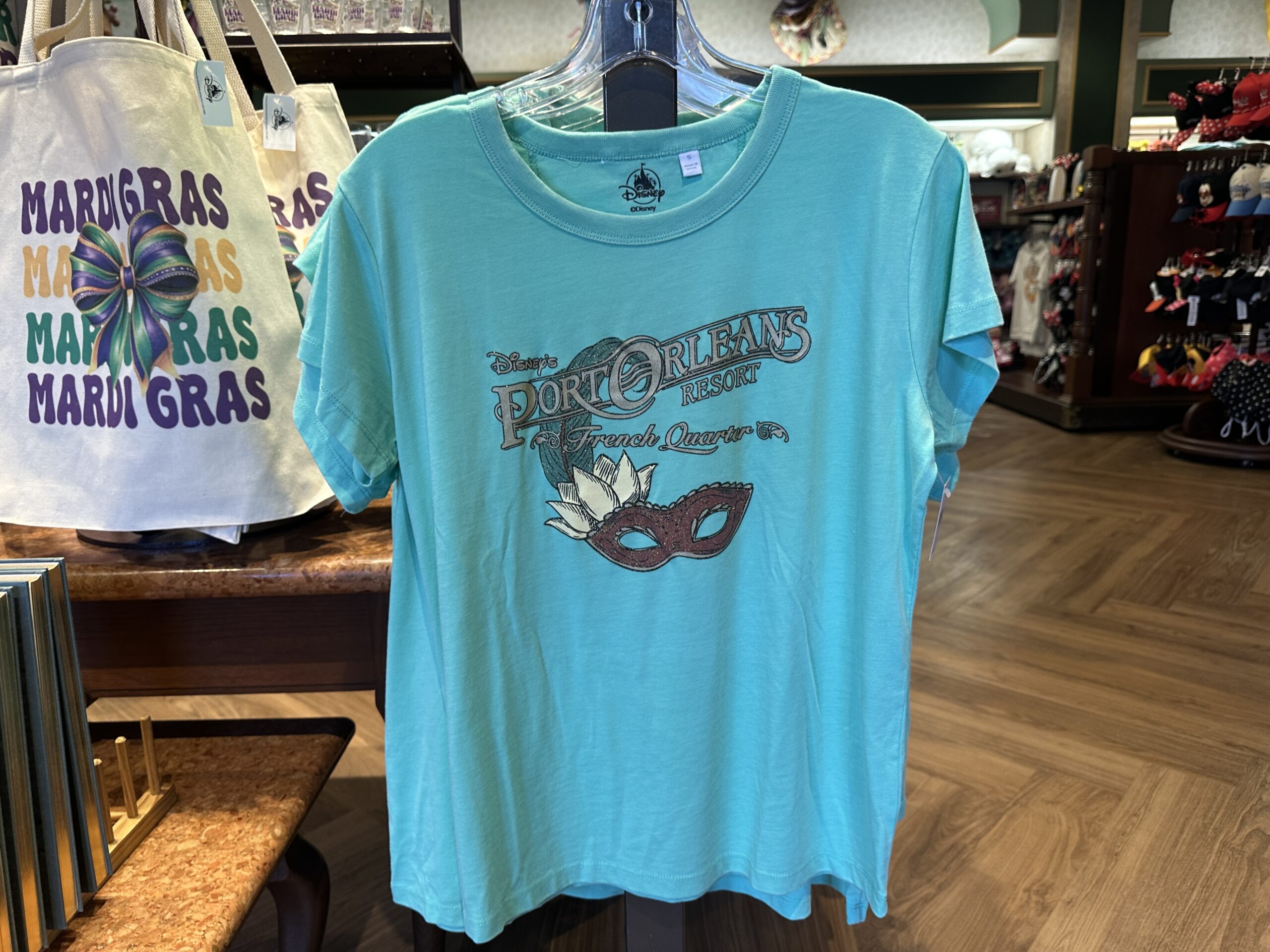 A sparkly purple Mardi Gras mask adorned with a lilypad and feather is pictured on this light blue tee. The resort's name is in sparkly gray lettering above the mask.