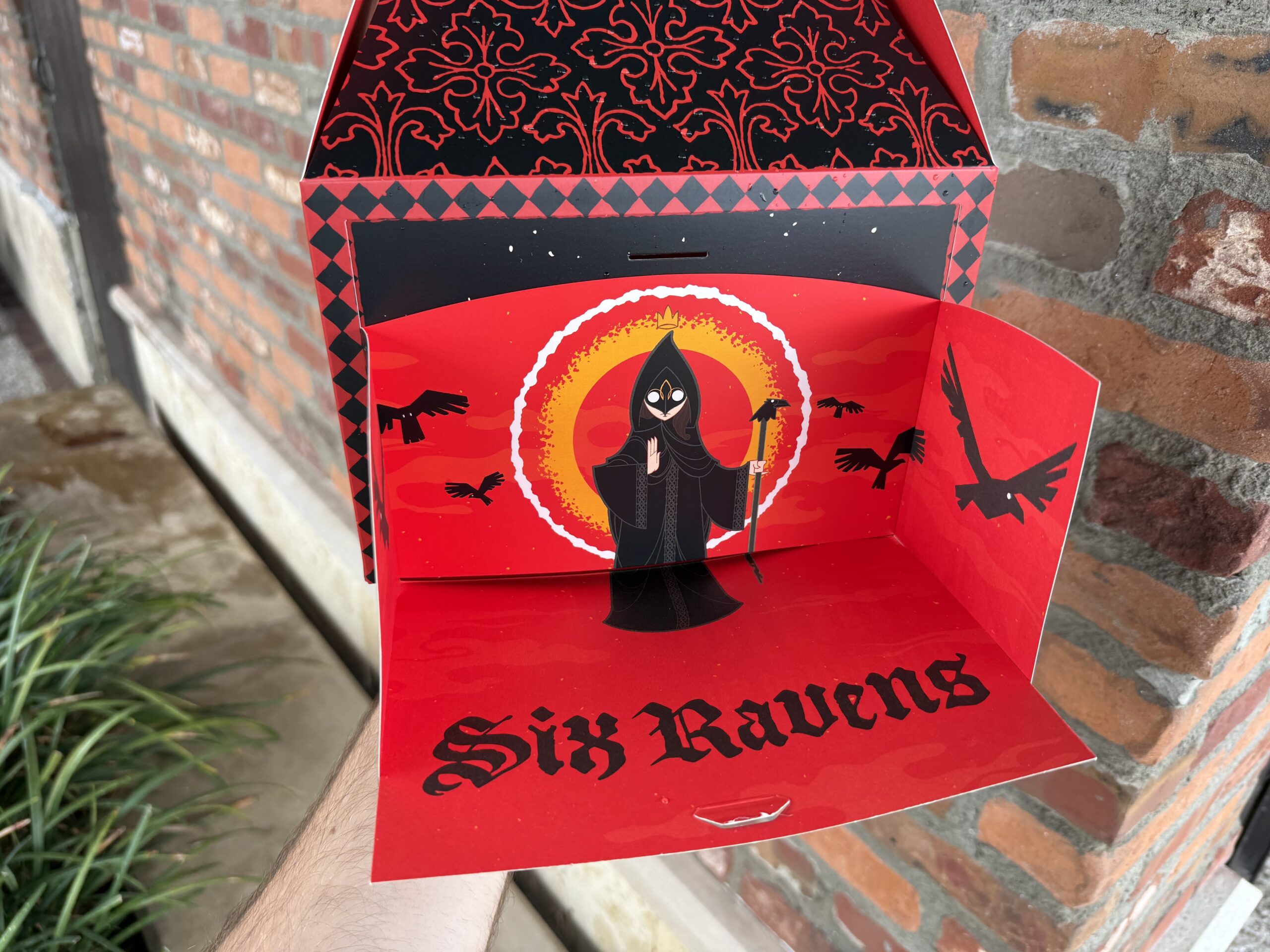 On one side is "Six Ravens" against a black background. On the reverse is the king from the restaurant's background story with "If it is good, 'tis better in a coffyn." That side opens up to create a kind of plate for eating on, featuring skeletal figures guarding a door and the six ravens with a cloaked figure.