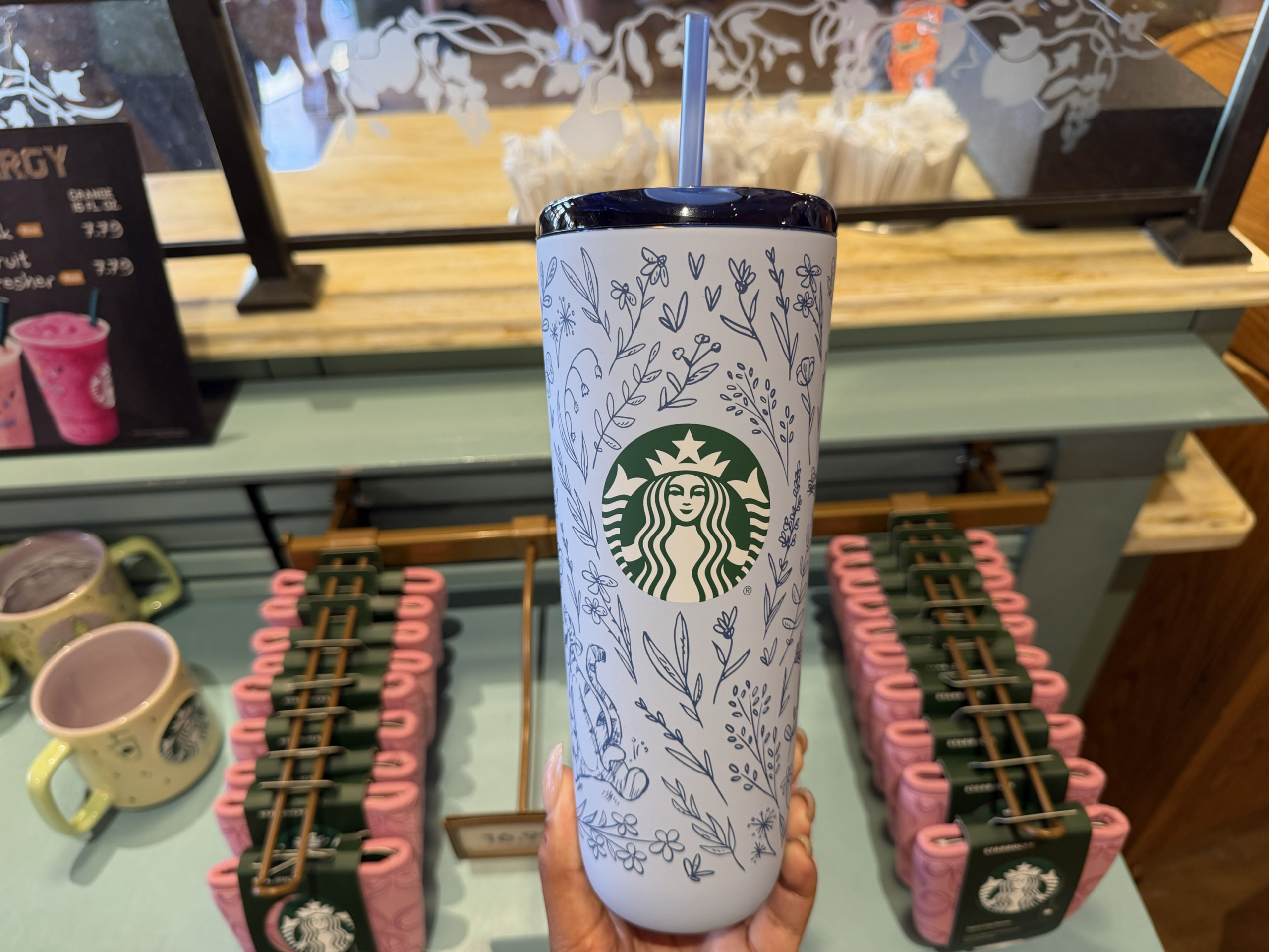 This light blue stainless steel tumbler is covered in delicate dark blue line drawings of flowers, leaves, butterflies, and characters from the Hundred Acre Wood, including Winnie the Pooh, Piglet, Tigger, and Eeyore. The green Starbucks logo sits on the front. It comes with a dark blue lid and matching straw.