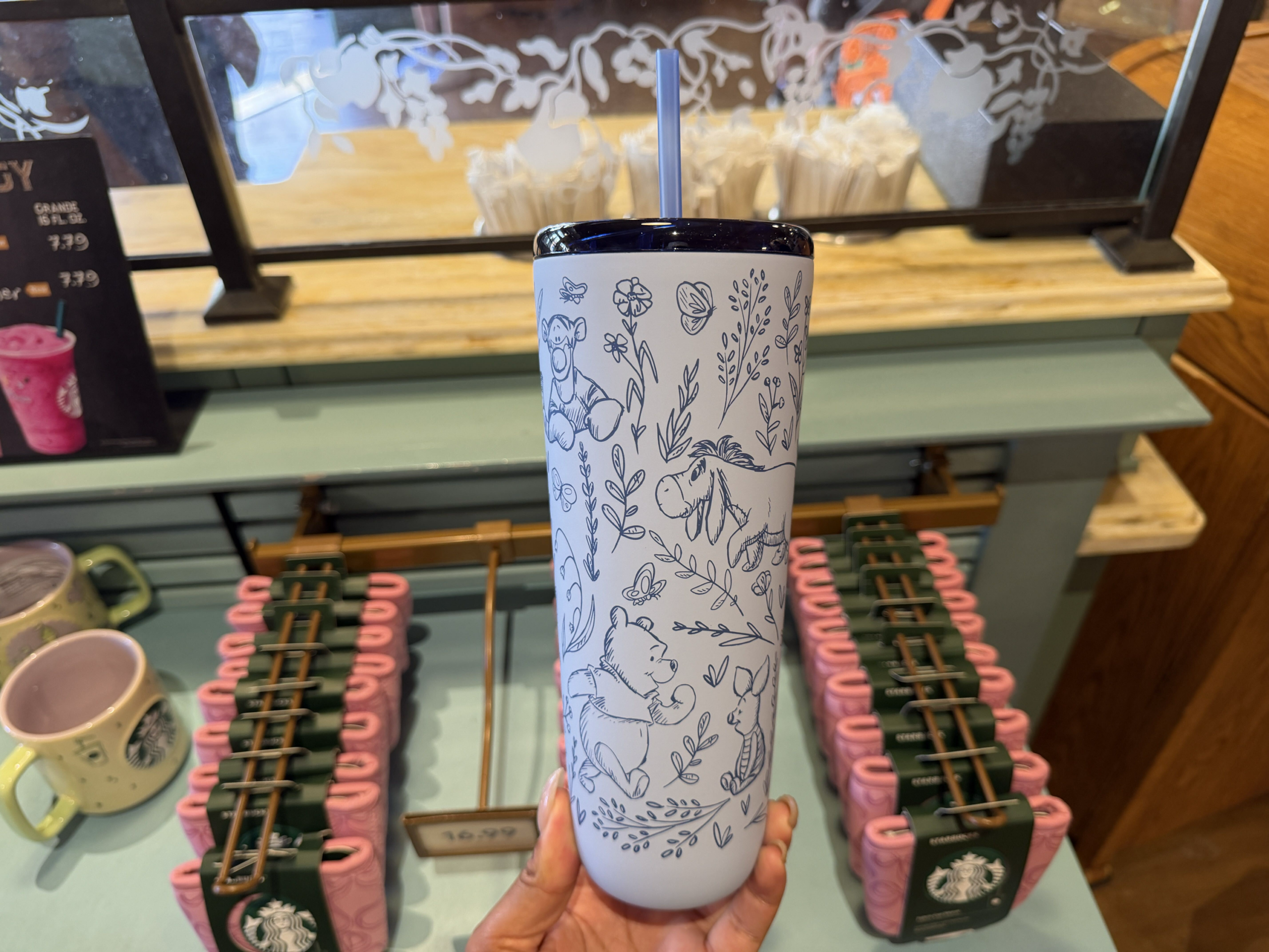 This light blue stainless steel tumbler is covered in delicate dark blue line drawings of flowers, leaves, butterflies, and characters from the Hundred Acre Wood, including Winnie the Pooh, Piglet, Tigger, and Eeyore. The green Starbucks logo sits on the front. It comes with a dark blue lid and matching straw.