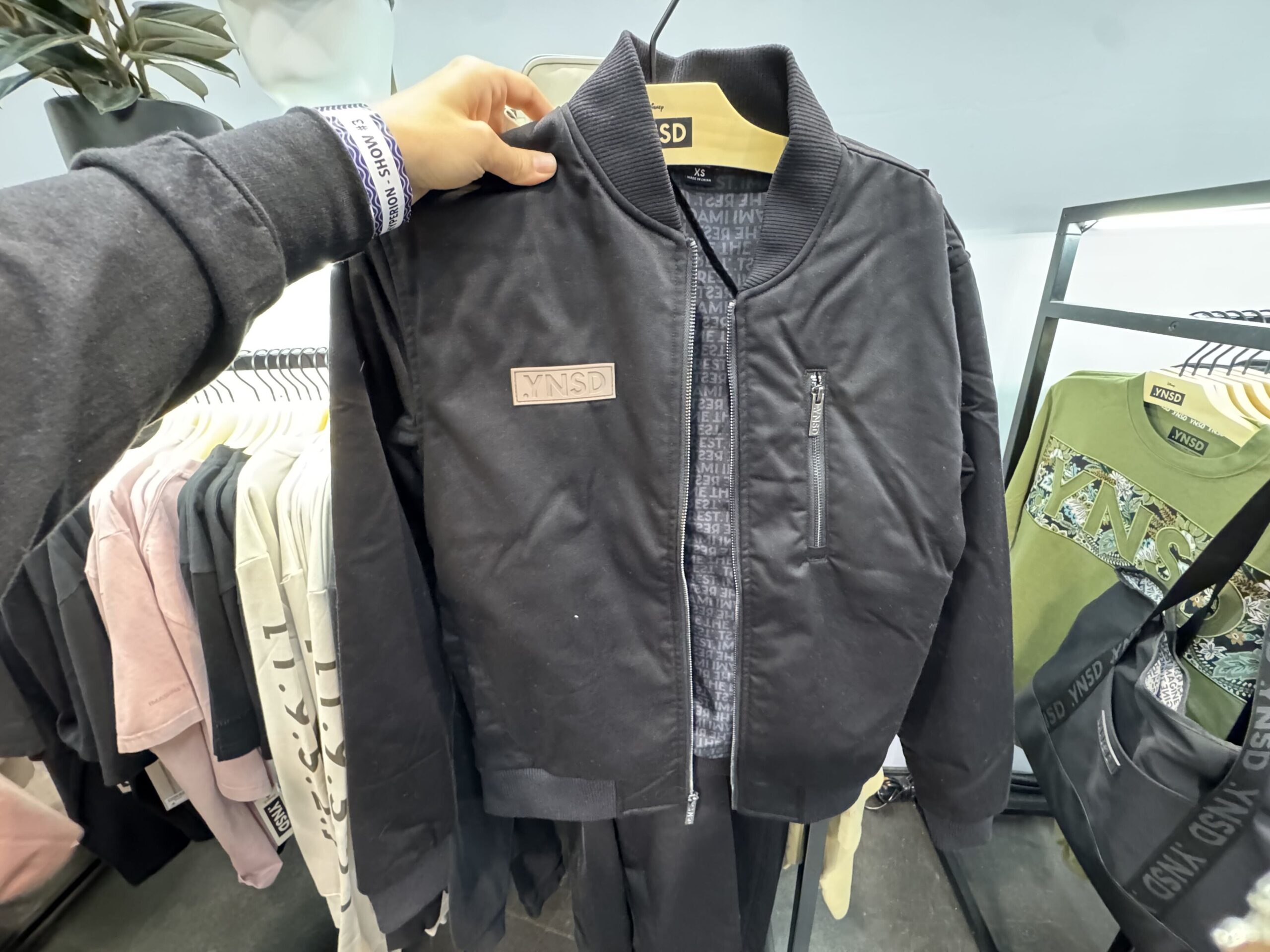 The black bomber jacket has the logo patch on the chest. "Imagine the Rest" is printed on the interior lining in a repeating pattern.