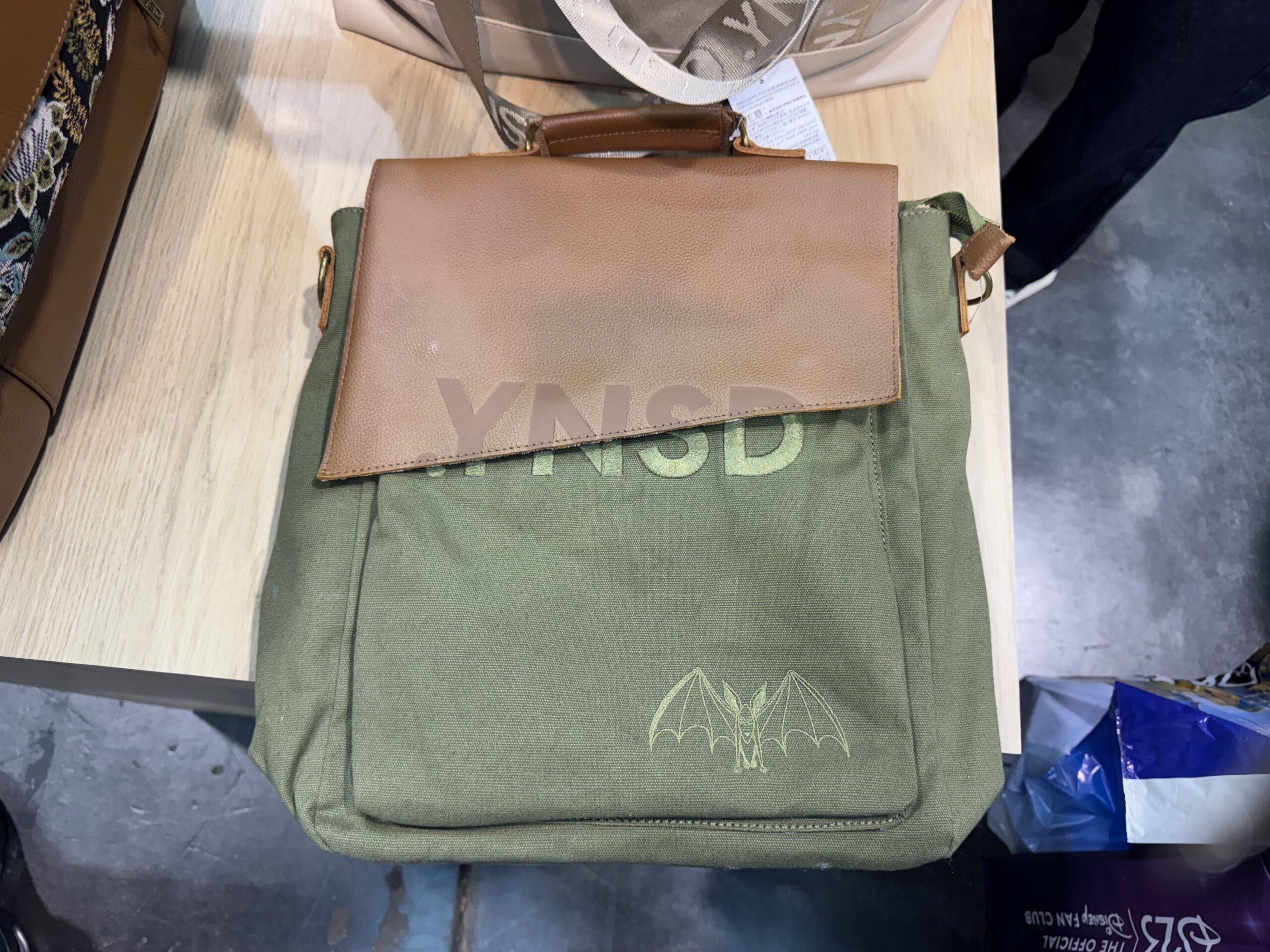 A convertible backpack is olive green and brown on one side (with the logo and Haunted Mansion spider) and carpet print on the other.