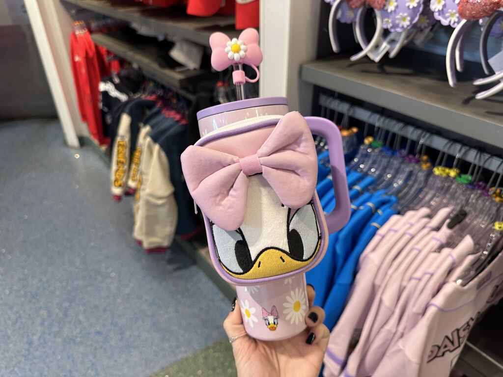 Daisy is featured on a light pink tumbler with white and yellow daisy flowers and a light purple lid and handle to match, adorned with a soft pouch featuring a close-up of Daisy's face and her signature pink bow. The bottom of the tumbler displays an icon of Daisy's's face, and the straw is topped with a silicone figure of her pink bow with a white and yellow daisy.