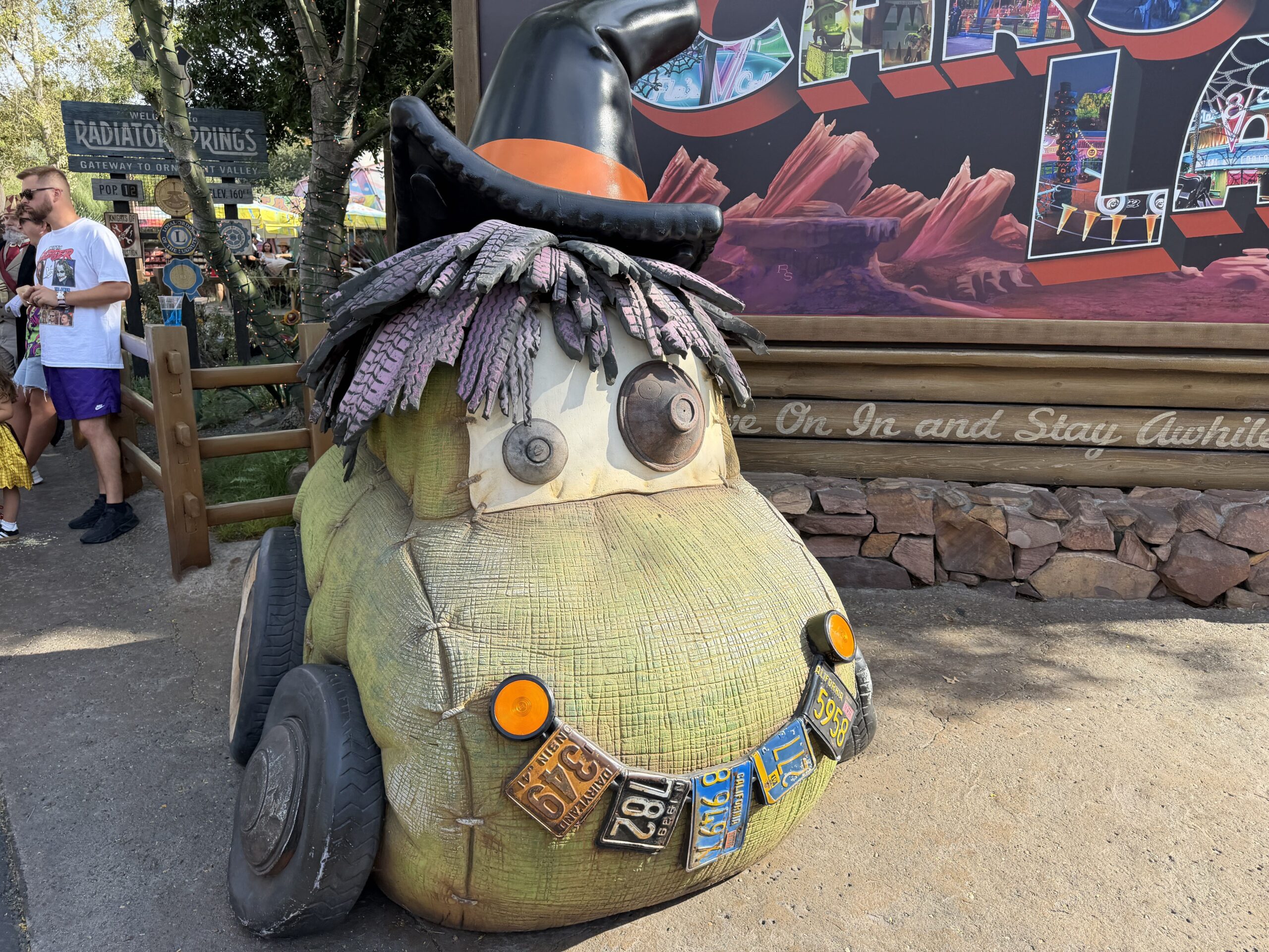 The ScareCar, wearing a black traffic cone as a witch's hat atop his mop of purple car wash brush hair, greets guests at the entrance of Cars Land, grinning a toothy, license plate smile.