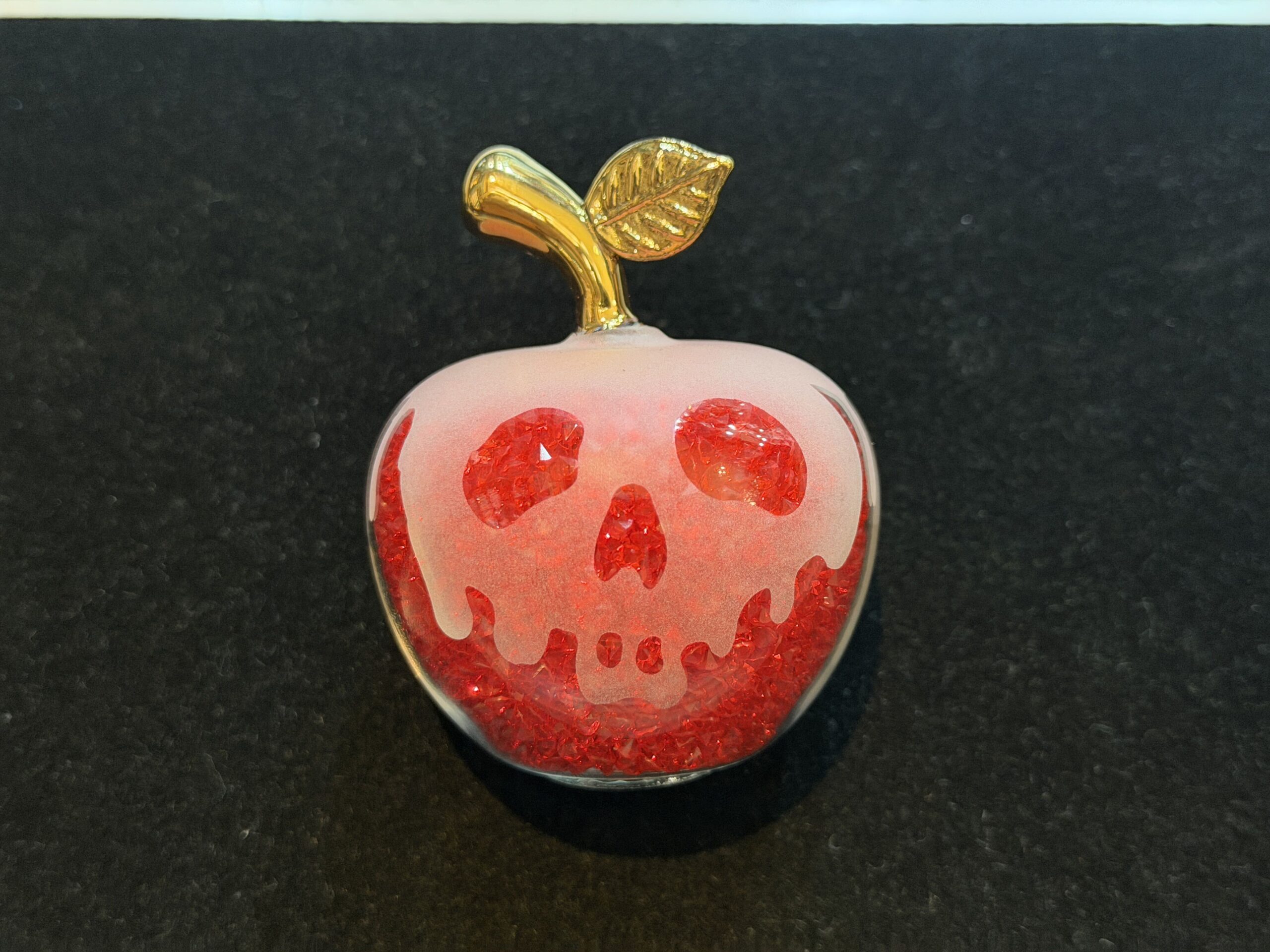 These tiny glass figures are miniatures of the poison apple that the Evil Queen, disguised as the Old Hag, offers to Snow White. One sparkles red and one sparkles black, and each has a dripping skeleton face, as seen on the poisoned apple in Snow White and the Seven Dwarves.
A short, gold stem with a single leaf tops each apple.