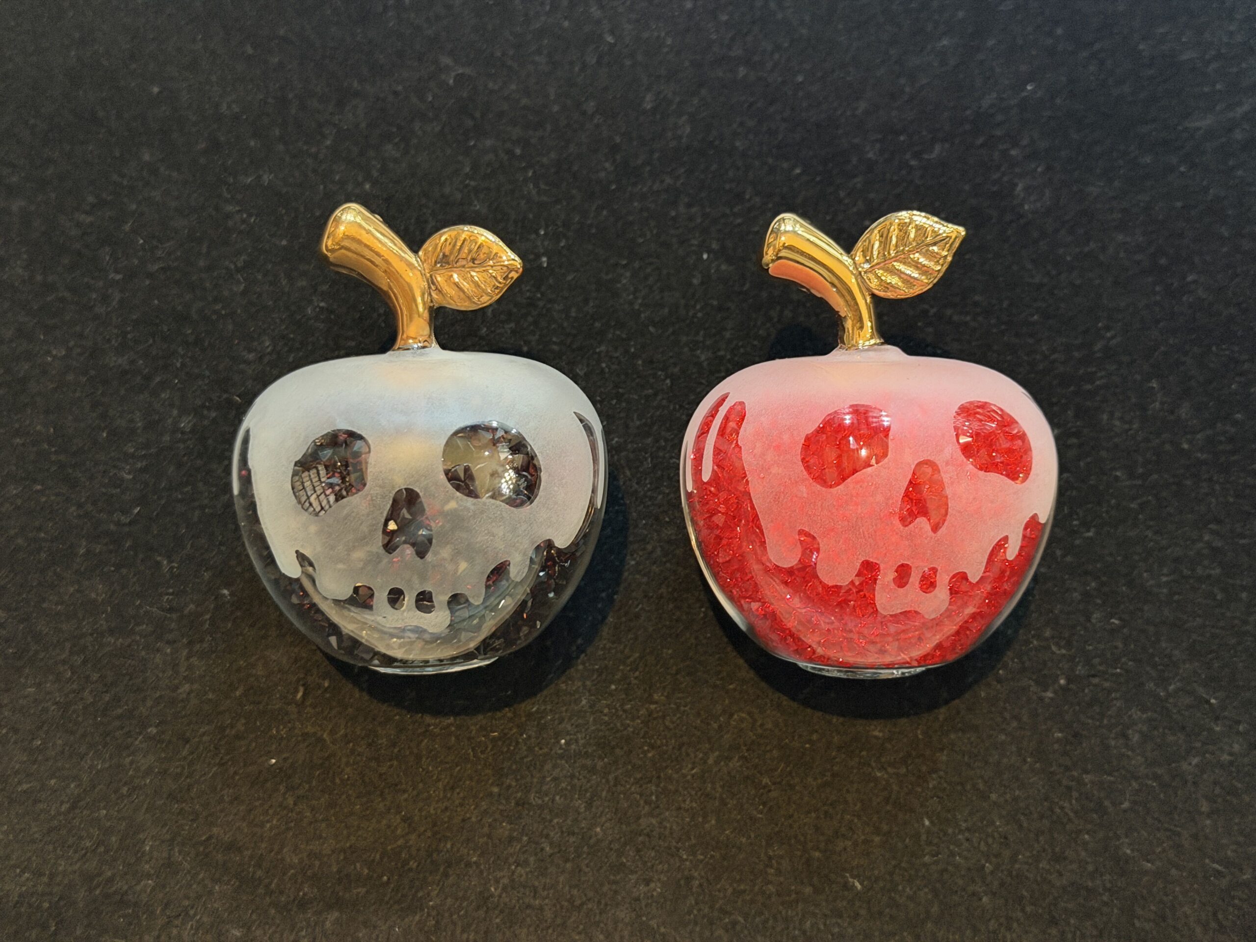 These tiny glass figures are miniatures of the poison apple that the Evil Queen, disguised as the Old Hag, offers to Snow White. One sparkles red and one sparkles black, and each has a dripping skeleton face, as seen on the poisoned apple in Snow White and the Seven Dwarves.
A short, gold stem with a single leaf tops each apple.