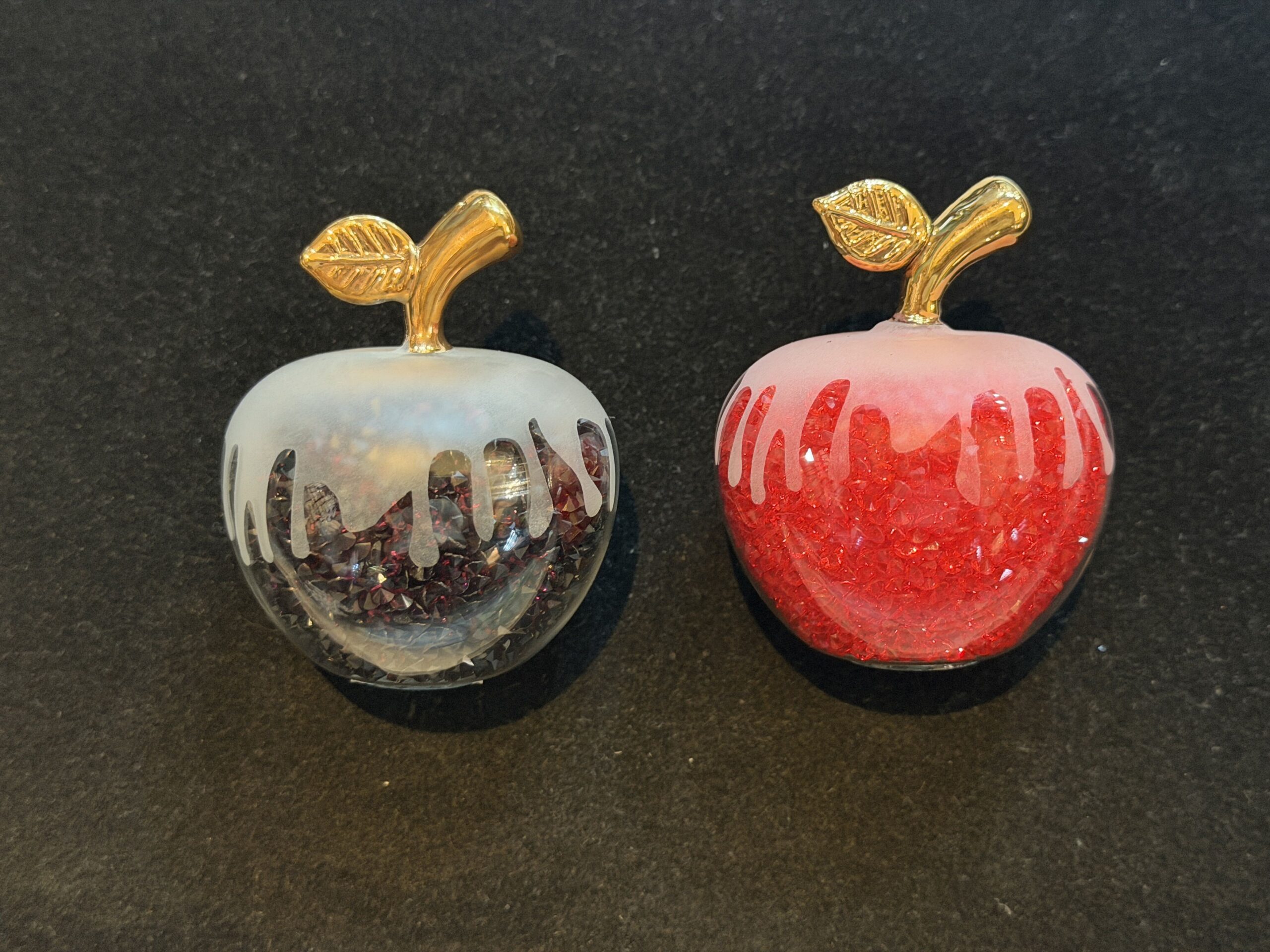 These tiny glass figures are miniatures of the poison apple that the Evil Queen, disguised as the Old Hag, offers to Snow White. One sparkles red and one sparkles black, and each has a dripping skeleton face, as seen on the poisoned apple in Snow White and the Seven Dwarves.
A short, gold stem with a single leaf tops each apple.
