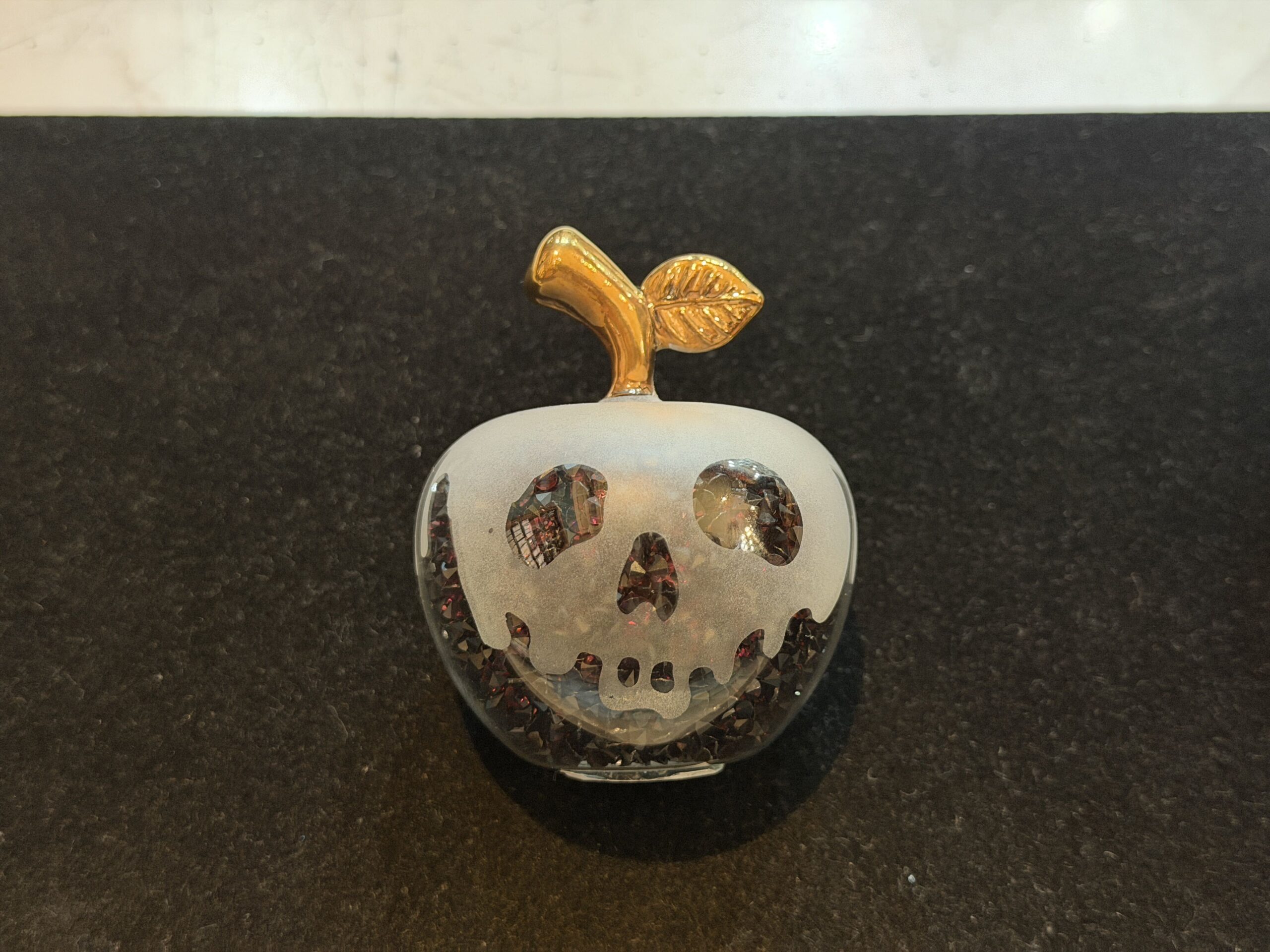 These tiny glass figures are miniatures of the poison apple that the Evil Queen, disguised as the Old Hag, offers to Snow White. One sparkles red and one sparkles black, and each has a dripping skeleton face, as seen on the poisoned apple in Snow White and the Seven Dwarves.
A short, gold stem with a single leaf tops each apple.