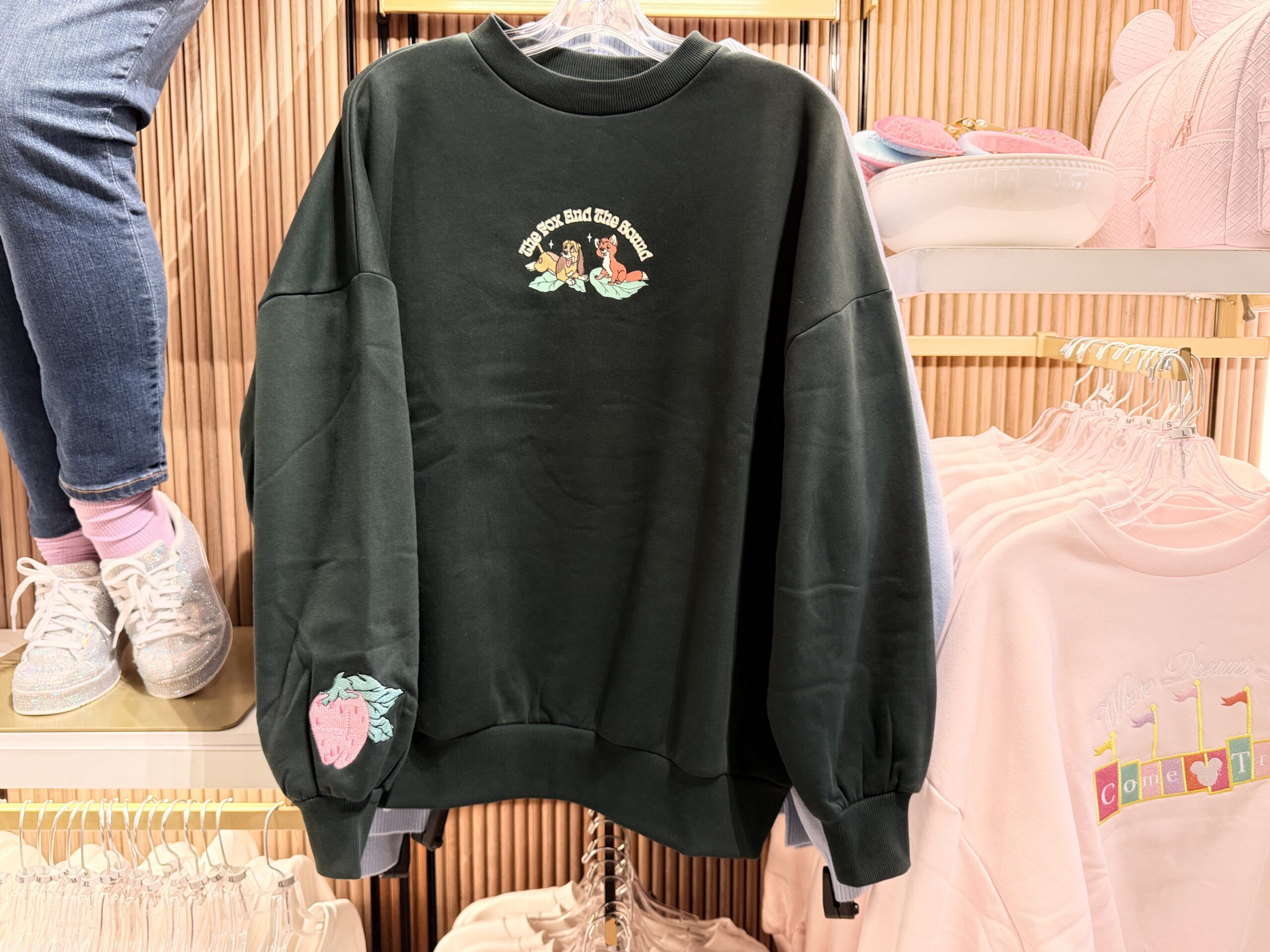 This black sweatshirt depicts Copper and Tod sitting upon green leaves. "The Fox and the Hound" is printed in yellow serif letters above the pair. A pink strawberry is embroidered on one sleeve end.