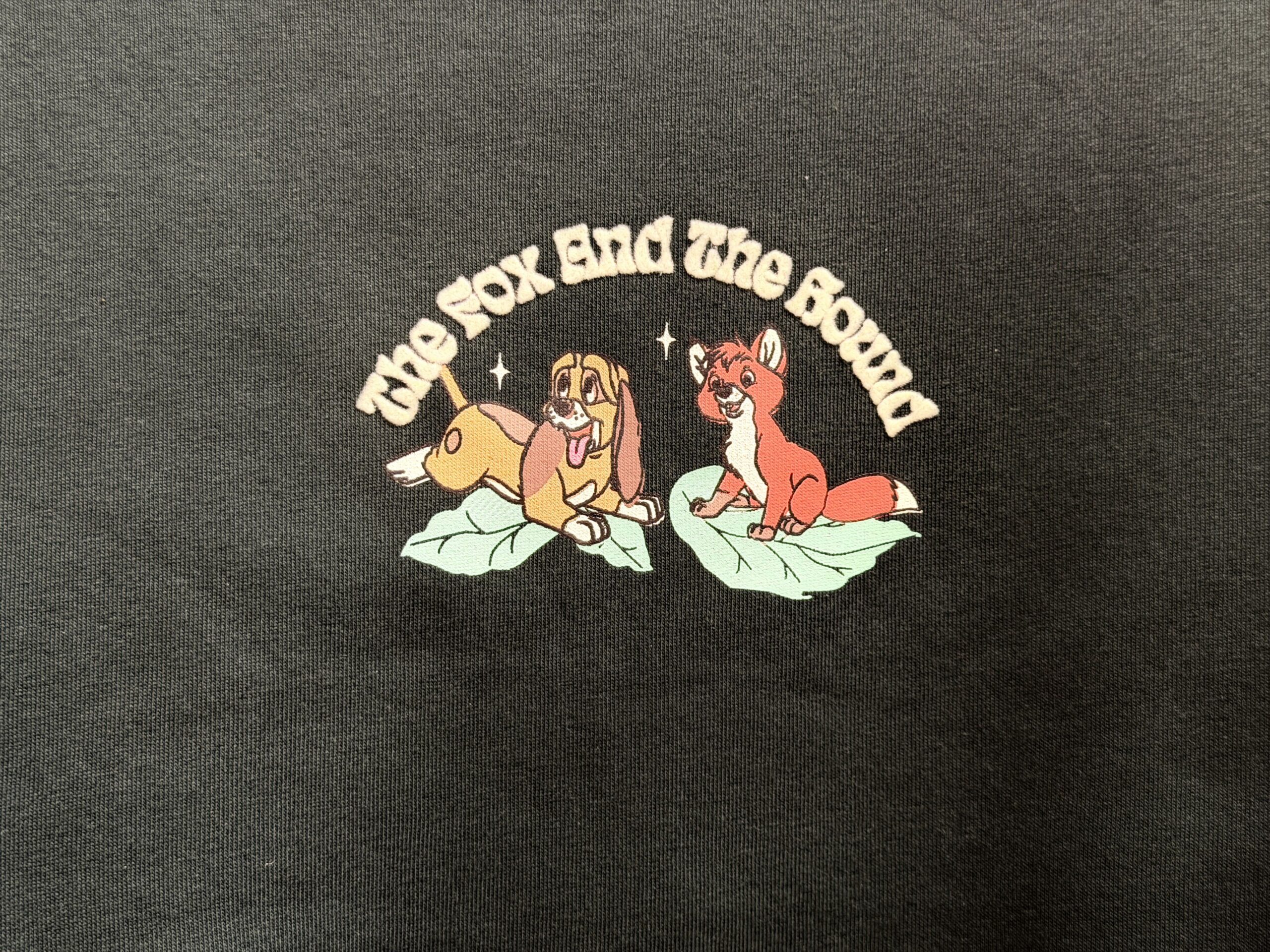 This black sweatshirt depicts Copper and Tod sitting upon green leaves. "The Fox and the Hound" is printed in yellow serif letters above the pair. A pink strawberry is embroidered on one sleeve end.