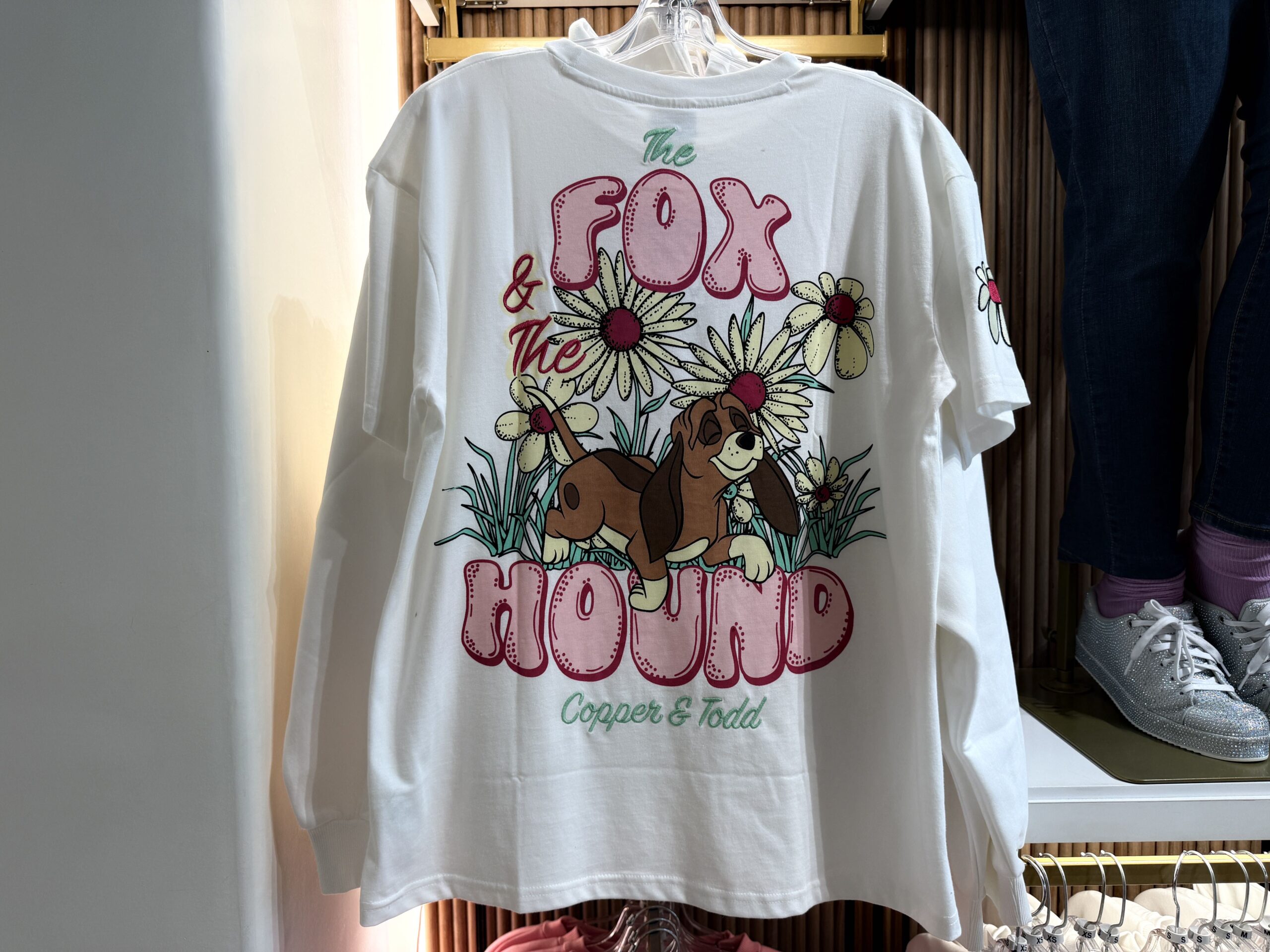 The back of the t-shirt depicts Copper the hound dog walking next to several tall white flowers. The "Fox" and "Hound" parts of the movie name are printed in large pink bubble letters. Tod's name is incorrectly spelled as "Todd".