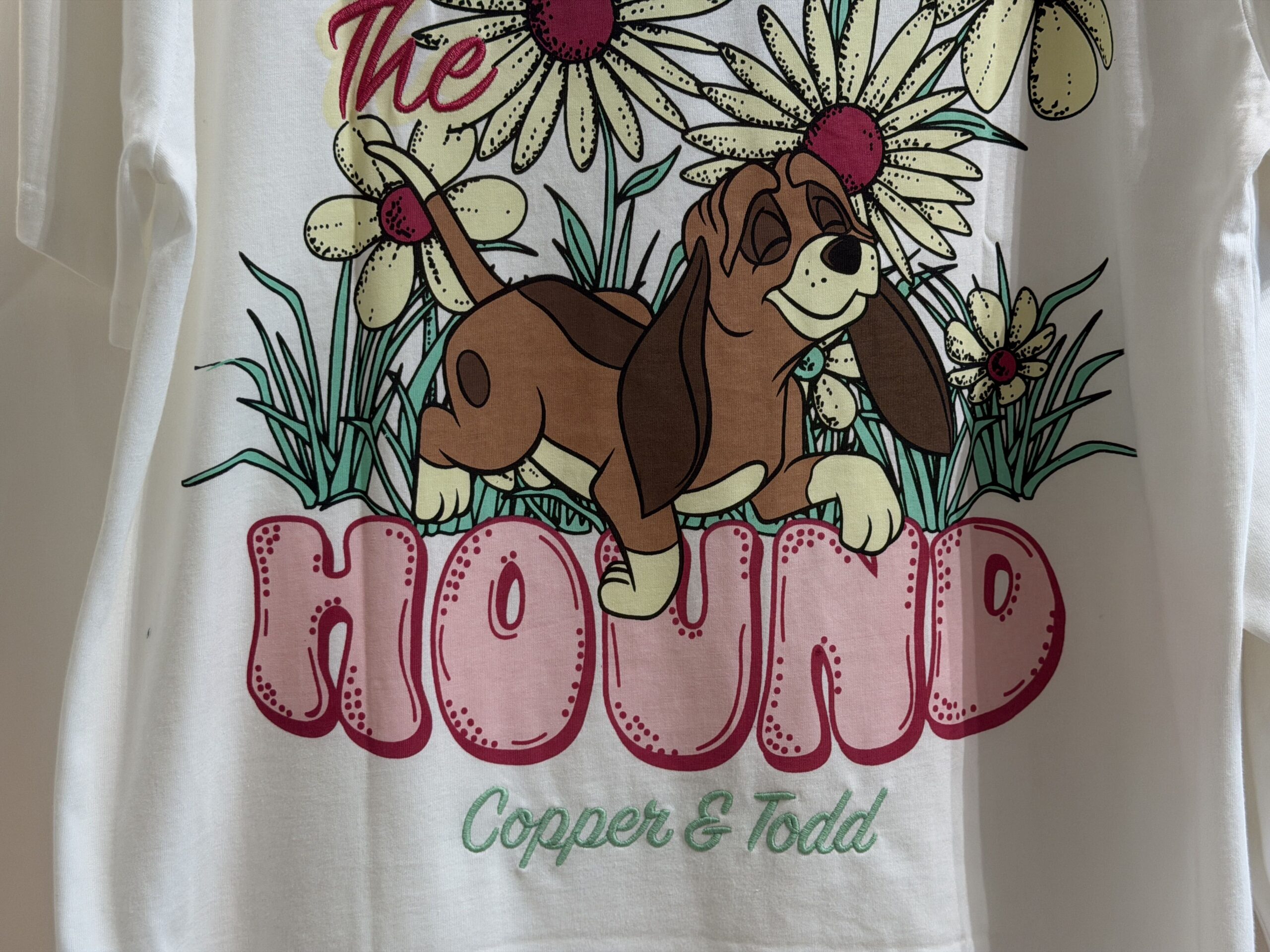 The back of the t-shirt depicts Copper the hound dog walking next to several tall white flowers. The "Fox" and "Hound" parts of the movie name are printed in large pink bubble letters. Tod's name is incorrectly spelled as "Todd".