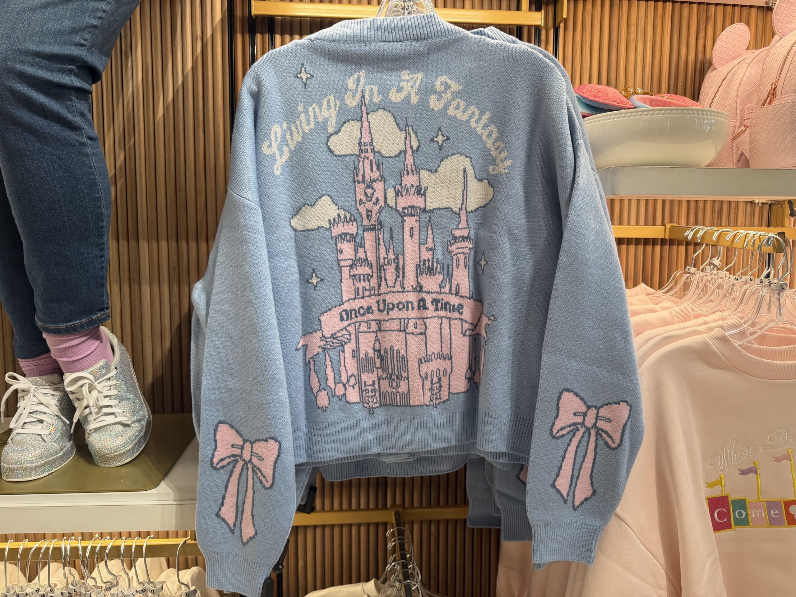 The back of the cardigan has a large pink Disney castle surrounded by white clouds and stars. "Living In A Fantasy" stretches in an arc above, while the text "Once Upon a Time" is shown on a pink scroll overlaying the castle.
