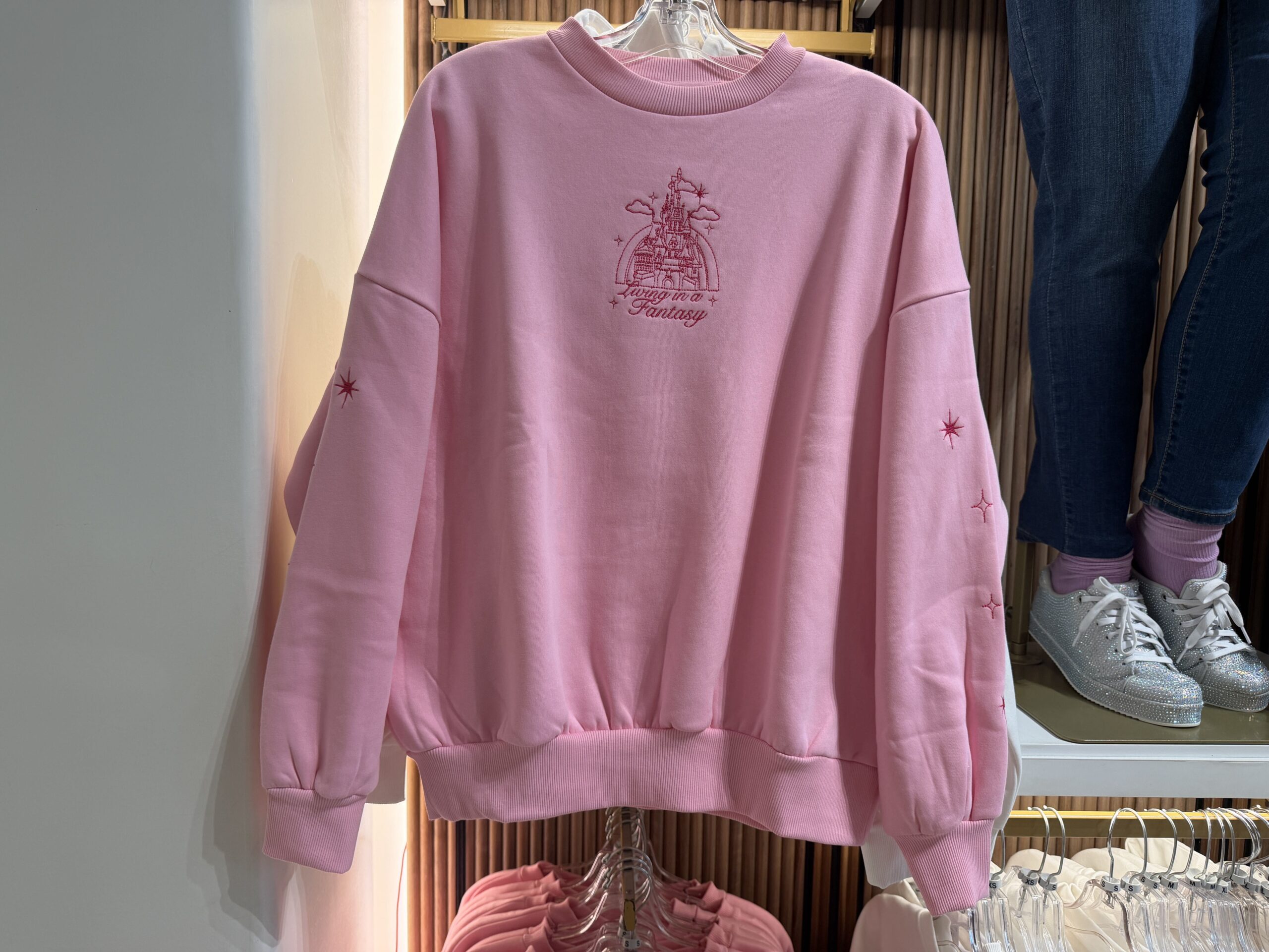 This pullover pink sweatshirt has a small front design consisting of a Disney castle embroidered in pink. The castle is surrounded by clouds and stars, and "Living in a fantasy" is embroidered in cursive below.
