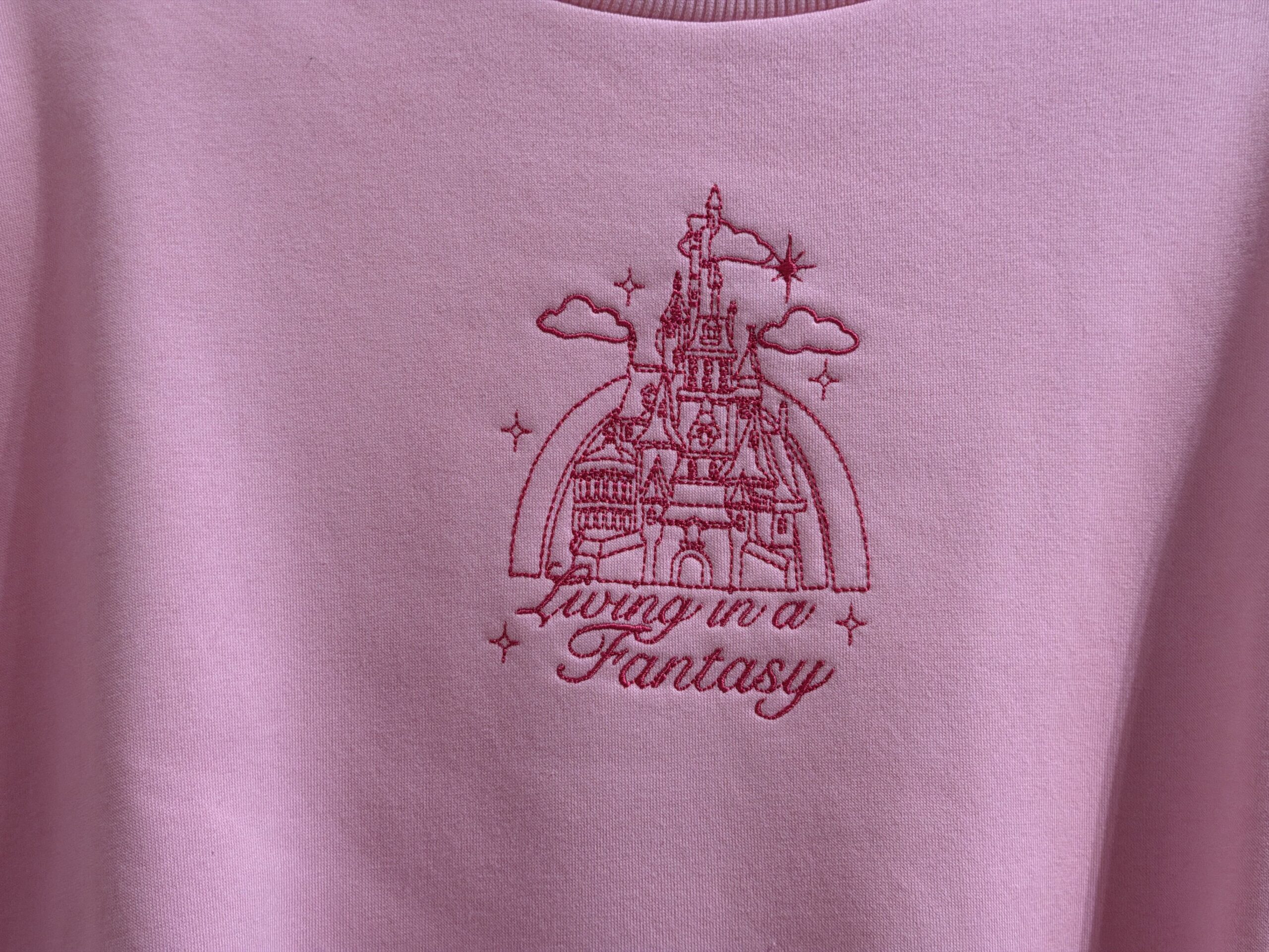 This pullover pink sweatshirt has a small front design consisting of a Disney castle embroidered in pink. The castle is surrounded by clouds and stars, and "Living in a fantasy" is embroidered in cursive below.