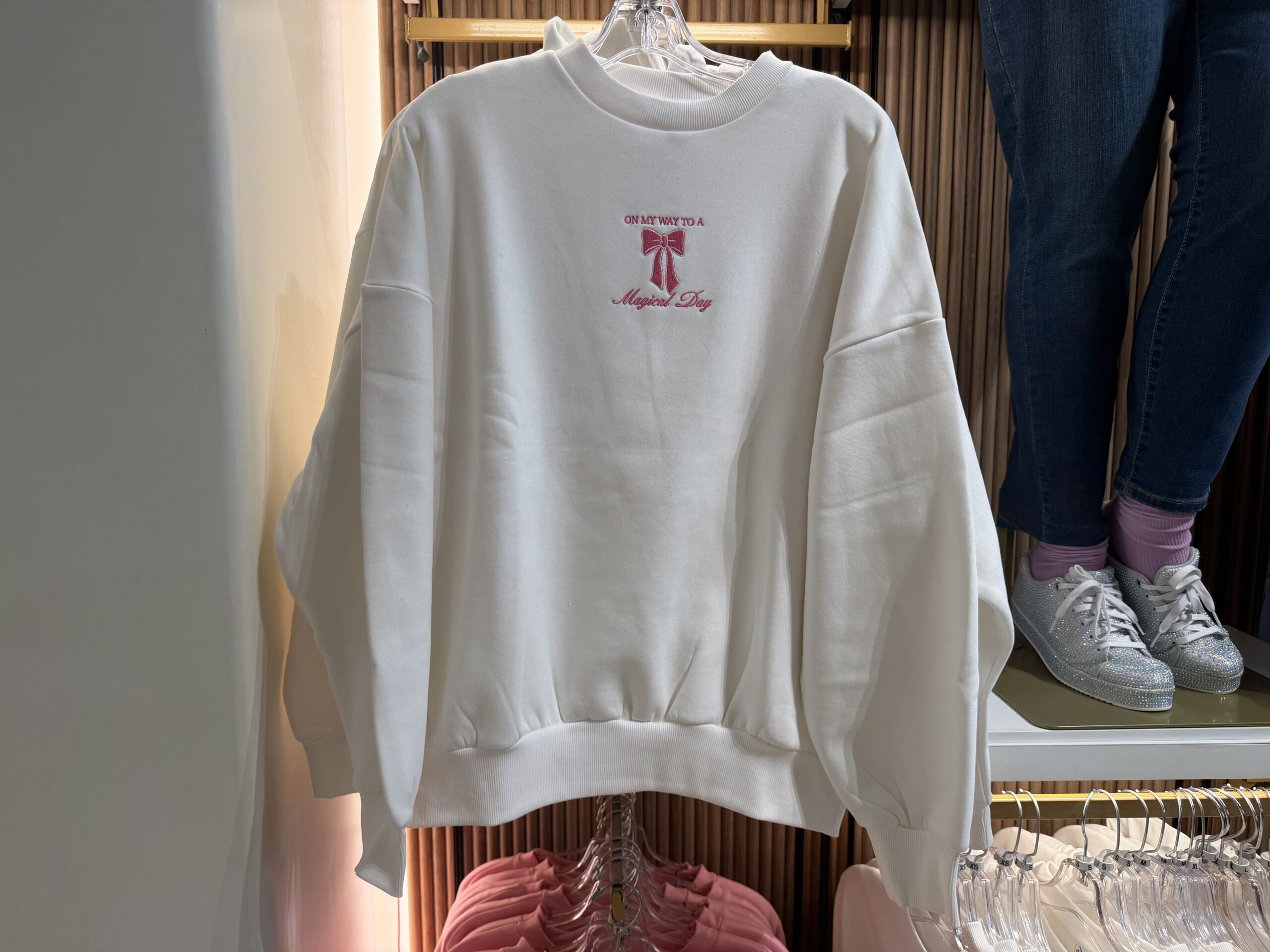 On this white sweatshirt, "On My Way To A Magical Day" is embroidered in pink thread. A small pink bow is placed in the middle of the text.