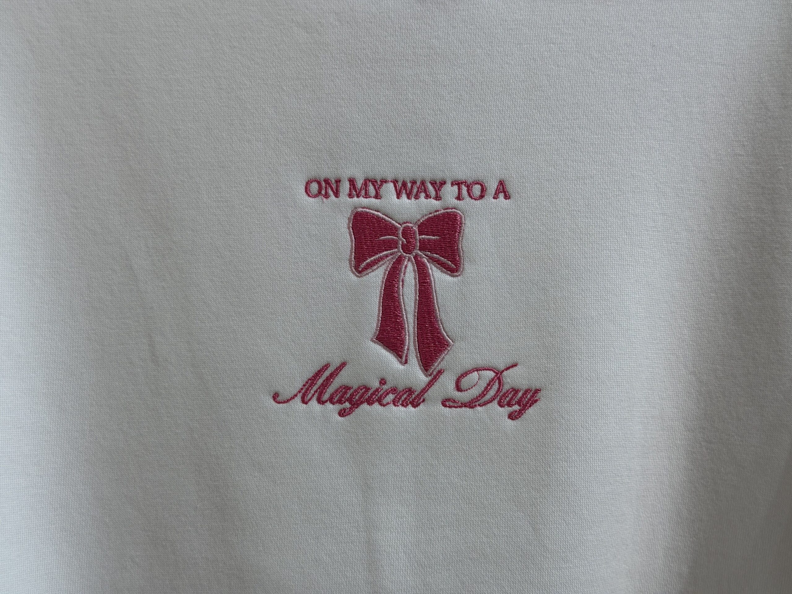 On this white sweatshirt, "On My Way To A Magical Day" is embroidered in pink thread. A small pink bow is placed in the middle of the text.