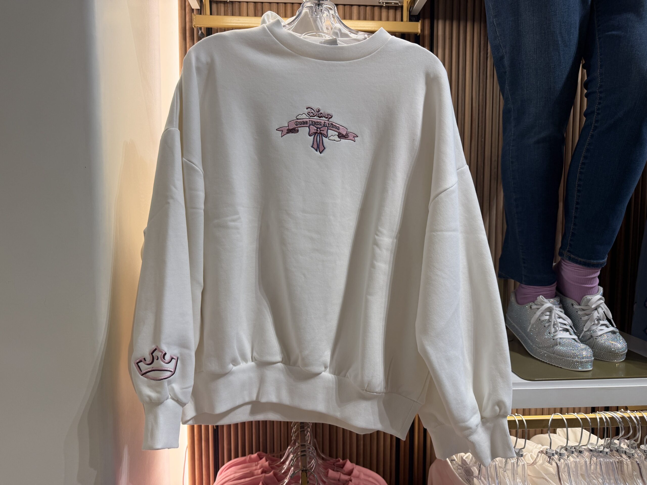 This white sweatshirt has "Once Upon a Time" embroidered on a pink scroll, surrounded by clouds and a pink bow. A pink crown edged in black is embroidered on one sleeve end.