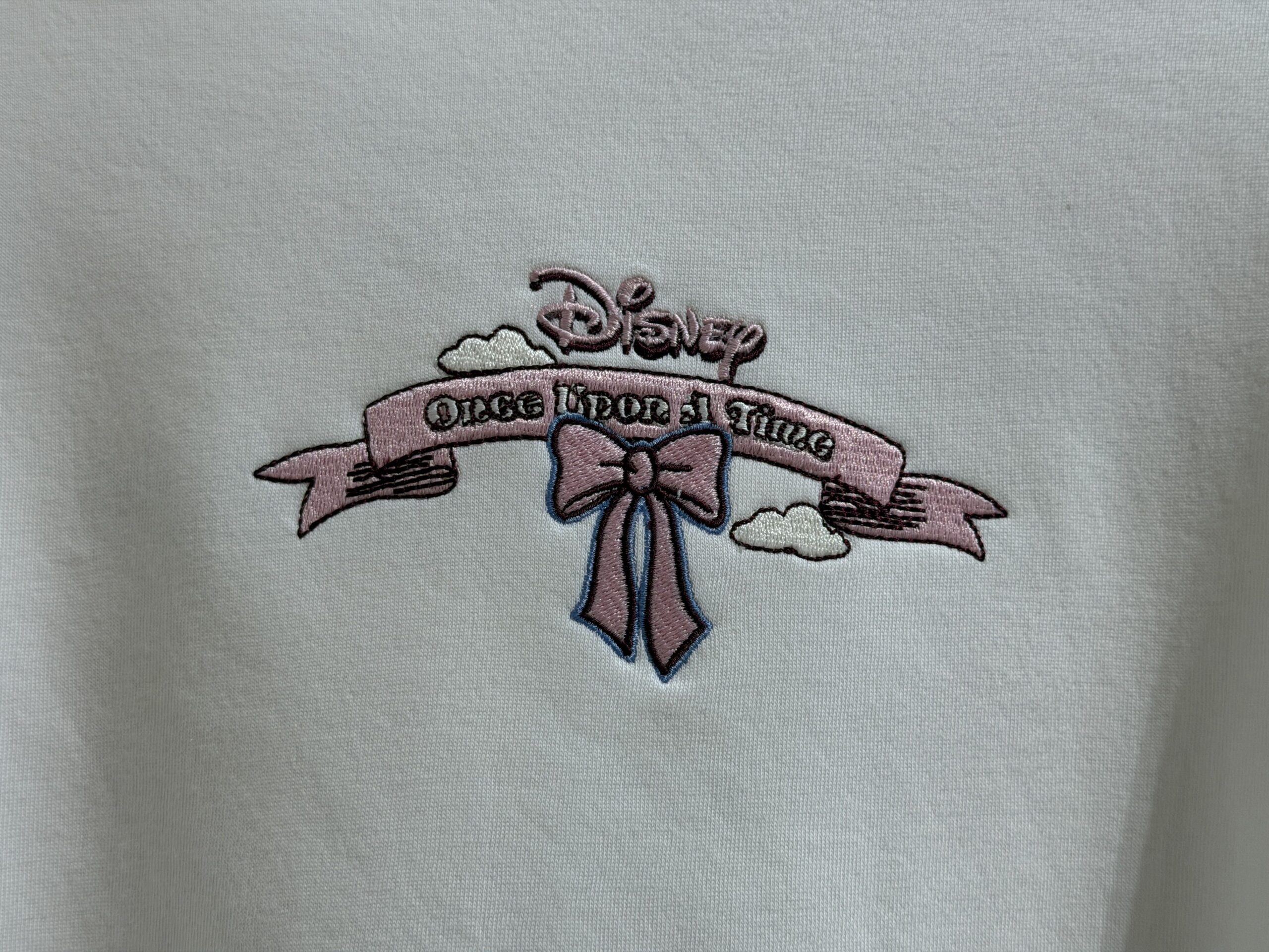 This white sweatshirt has "Once Upon a Time" embroidered on a pink scroll, surrounded by clouds and a pink bow. A pink crown edged in black is embroidered on one sleeve end.