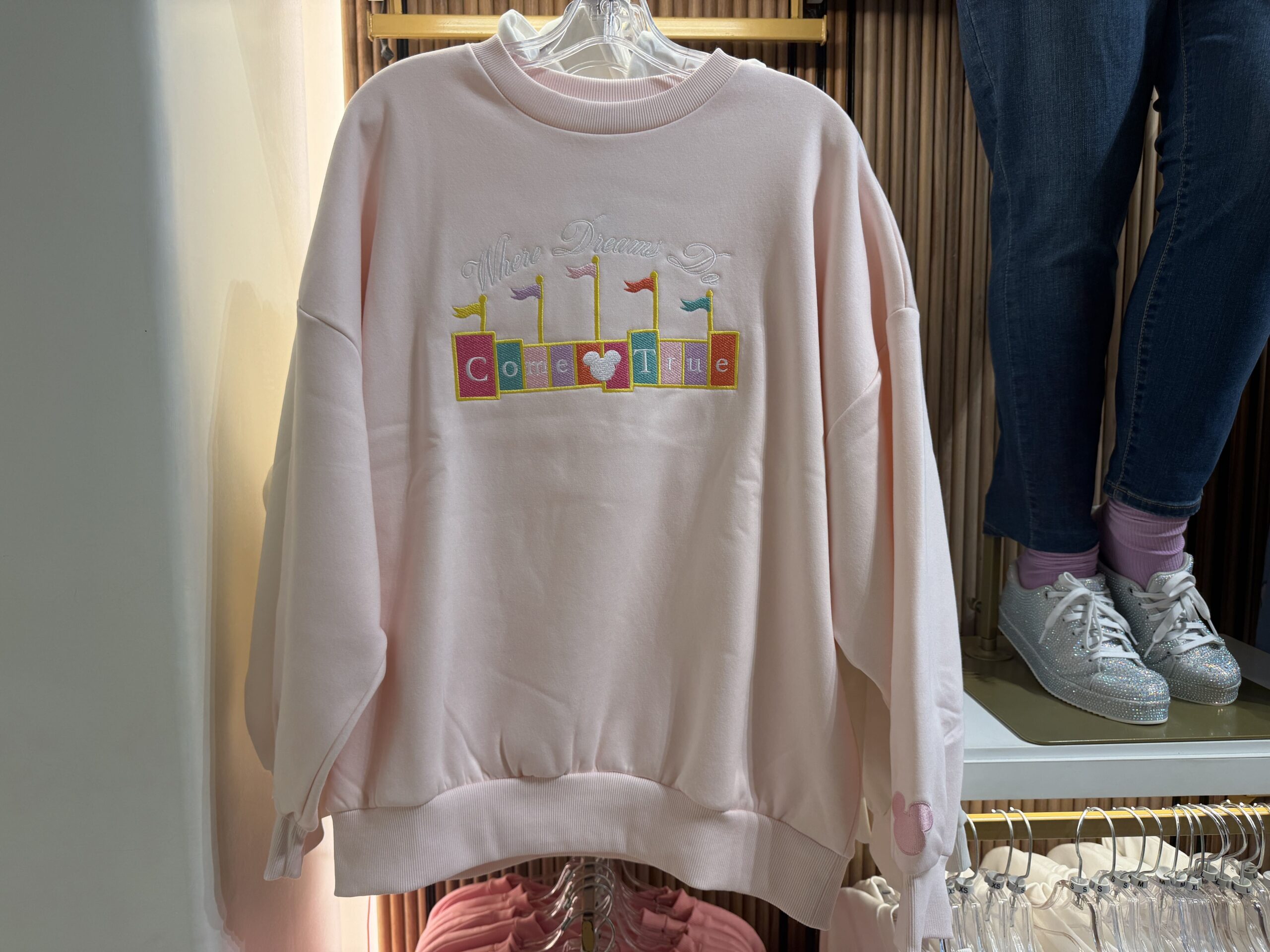This faintly pink sweatshirt has "Where Dreams Do" embroidered in white thread above a color block marquee sign. The sign completes the phrase by saying, "Come True." A white Mickey head is placed in the middle of the sign, and there are multicolored flags added on top.