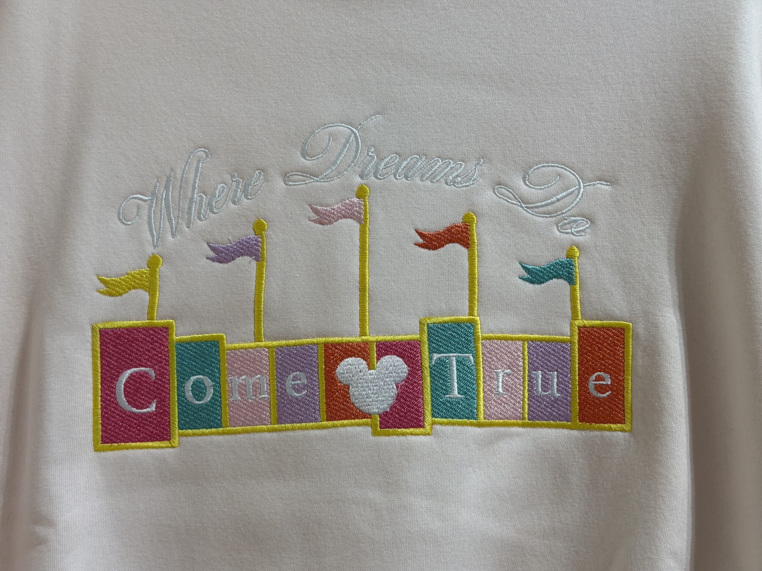 This faintly pink sweatshirt has "Where Dreams Do" embroidered in white thread above a color block marquee sign. The sign completes the phrase by saying, "Come True." A white Mickey head is placed in the middle of the sign, and there are multicolored flags added on top.