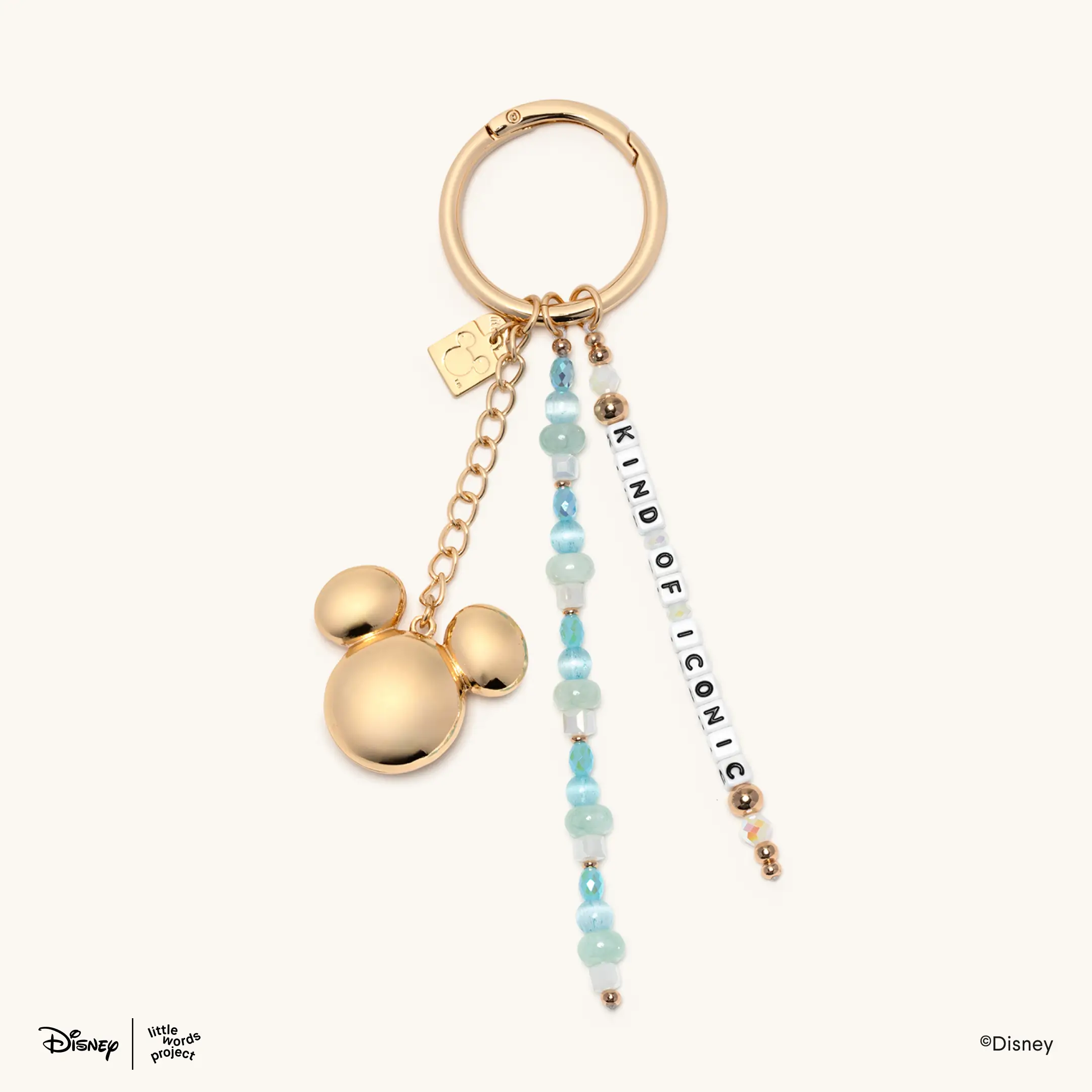 This bag charm has three items attached to a gold ring: A strand of gold and clear beads with "KIND OF ICONIC" printed on white letter blocks; a strand of white, sea foam green, and aqua beads; and a large gold Mickey head charm attached to a gold chain.