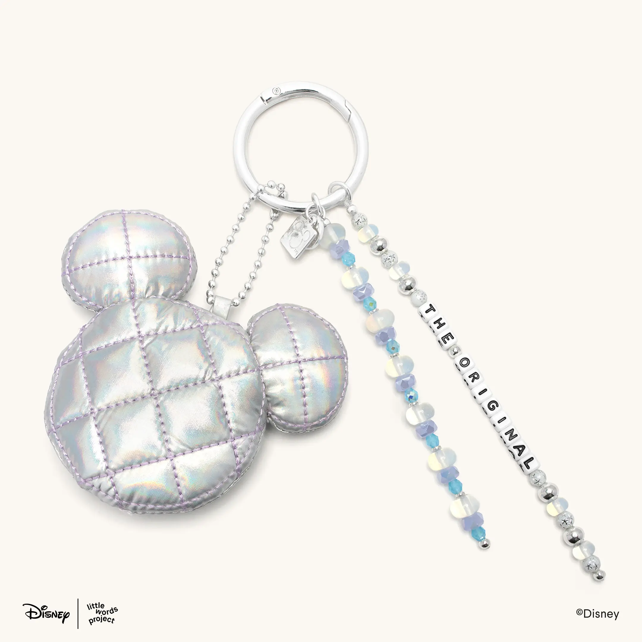 This bag charm has three items attached to a silver ring: A strand of silver and clear beads with "THE ORIGINAL" printed on white letter blocks; a strand of clear and blue beads; and an iridescent, puffy Mickey head charm.