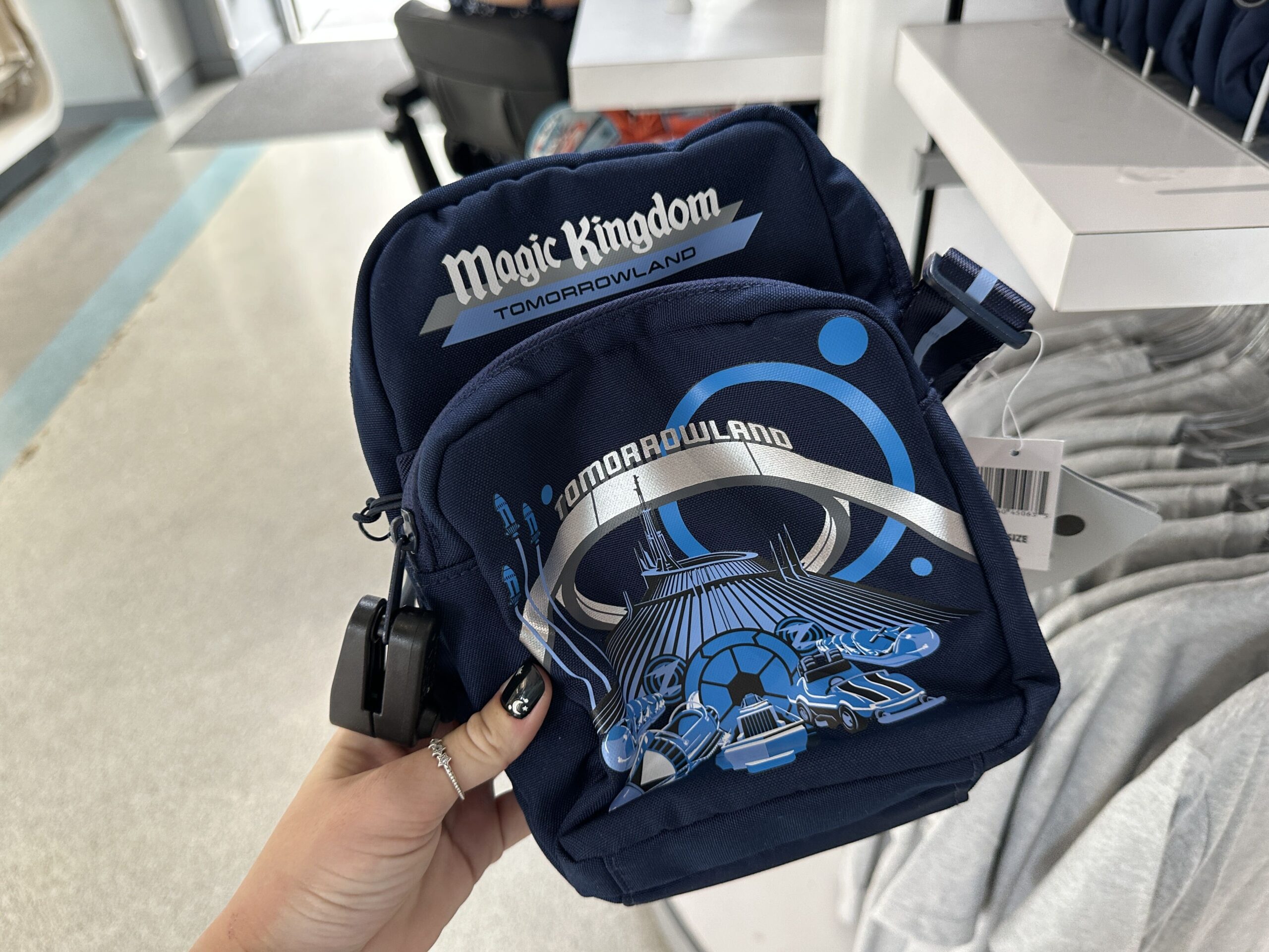 A Tomorrowland-themed crossbody bag debuts alongside the T-shirt to match. The bag is navy blue and has top and front zip pockets. On the top of the front zip pocket, "Magic Kingdom" appears in white screen print lettering. Below on the bottom front zip pocket is a screen-printed blue image of Space Mountain and the archway reading "Tomorrowland" in silver. The bag strap features a light blue Autopia car and road.