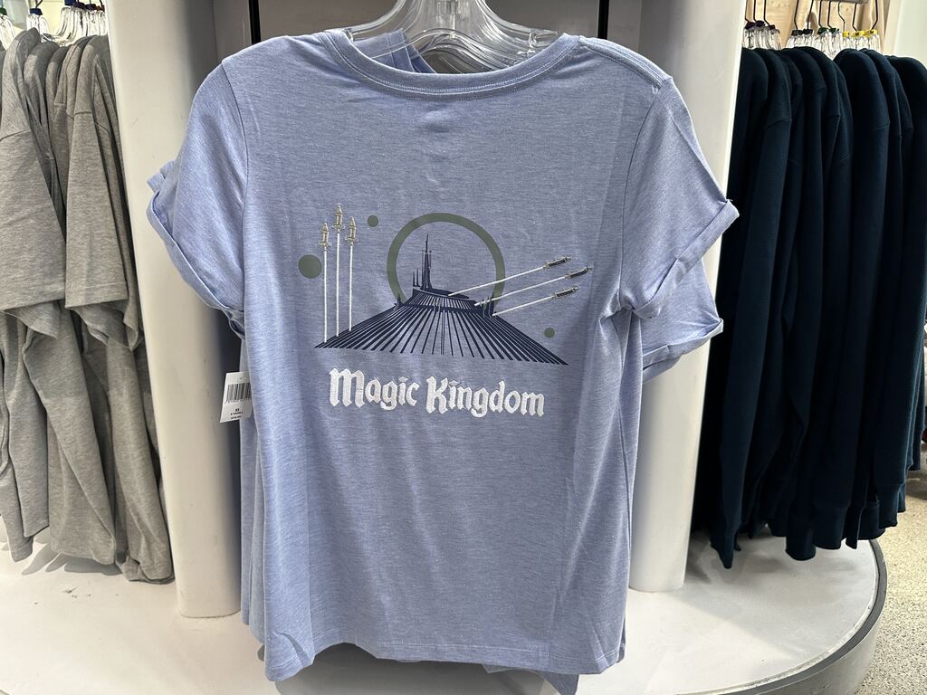 The Tomorrowland T-shirt is pale blue with sparkles, and features "Tomorrowland" in white embroidery across the front. The back of the shirt features Space Mountain in navy blue in the center, with "Magic Kingdom" embroidered in white below it. On either side of Space Mountain, there are planets and rocket ships embroidered in white, tan, and gray, launching to the sky on the left-hand side and diagonally to the right.