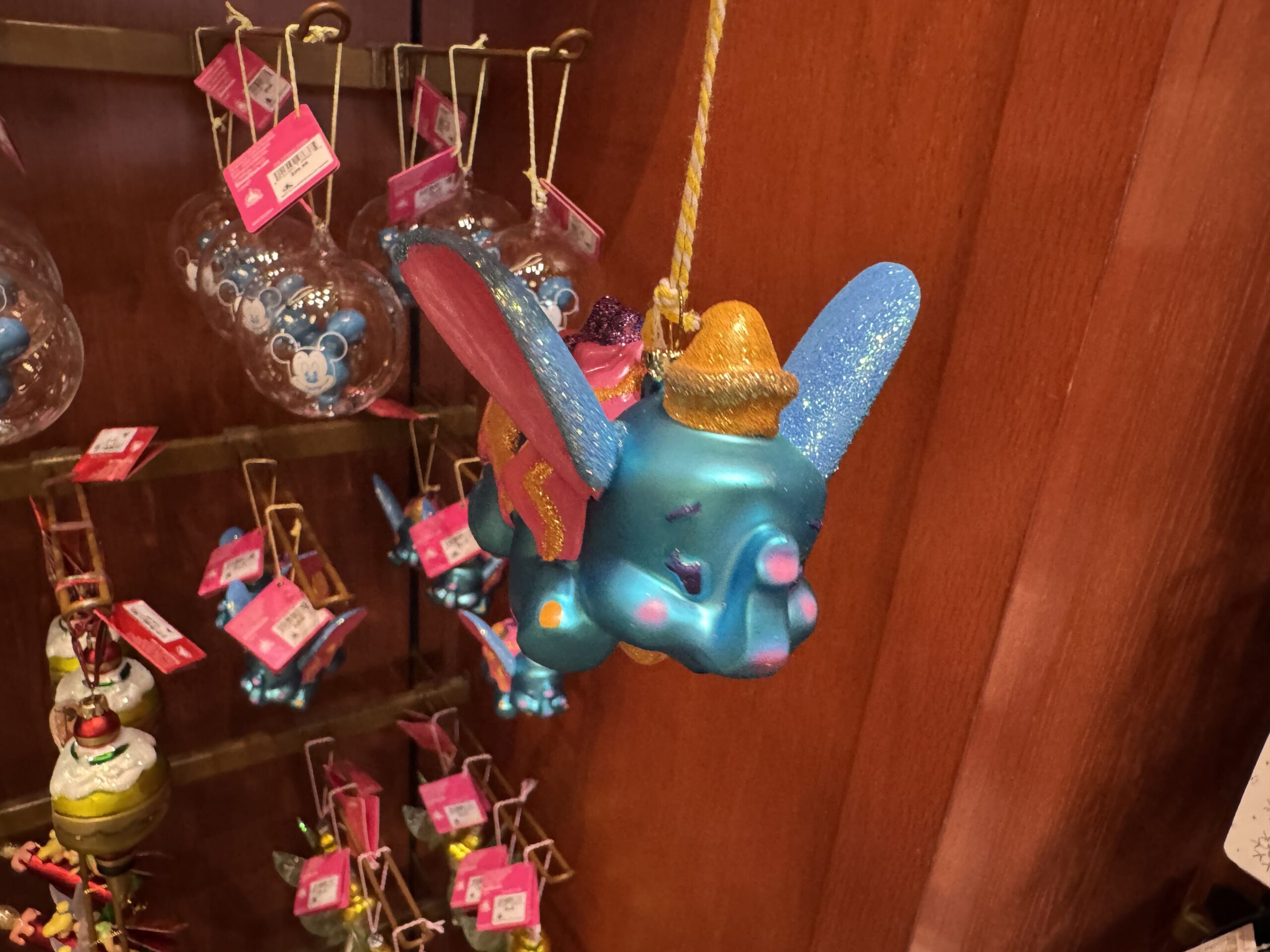 Dumbo flies in bright, cheery colors of blue, orange, and pink. His ears, hat, and saddle holding presents sparkle with glitter. He is crafted of porcelain and painted with rosy cheeks in a watercolor style. 