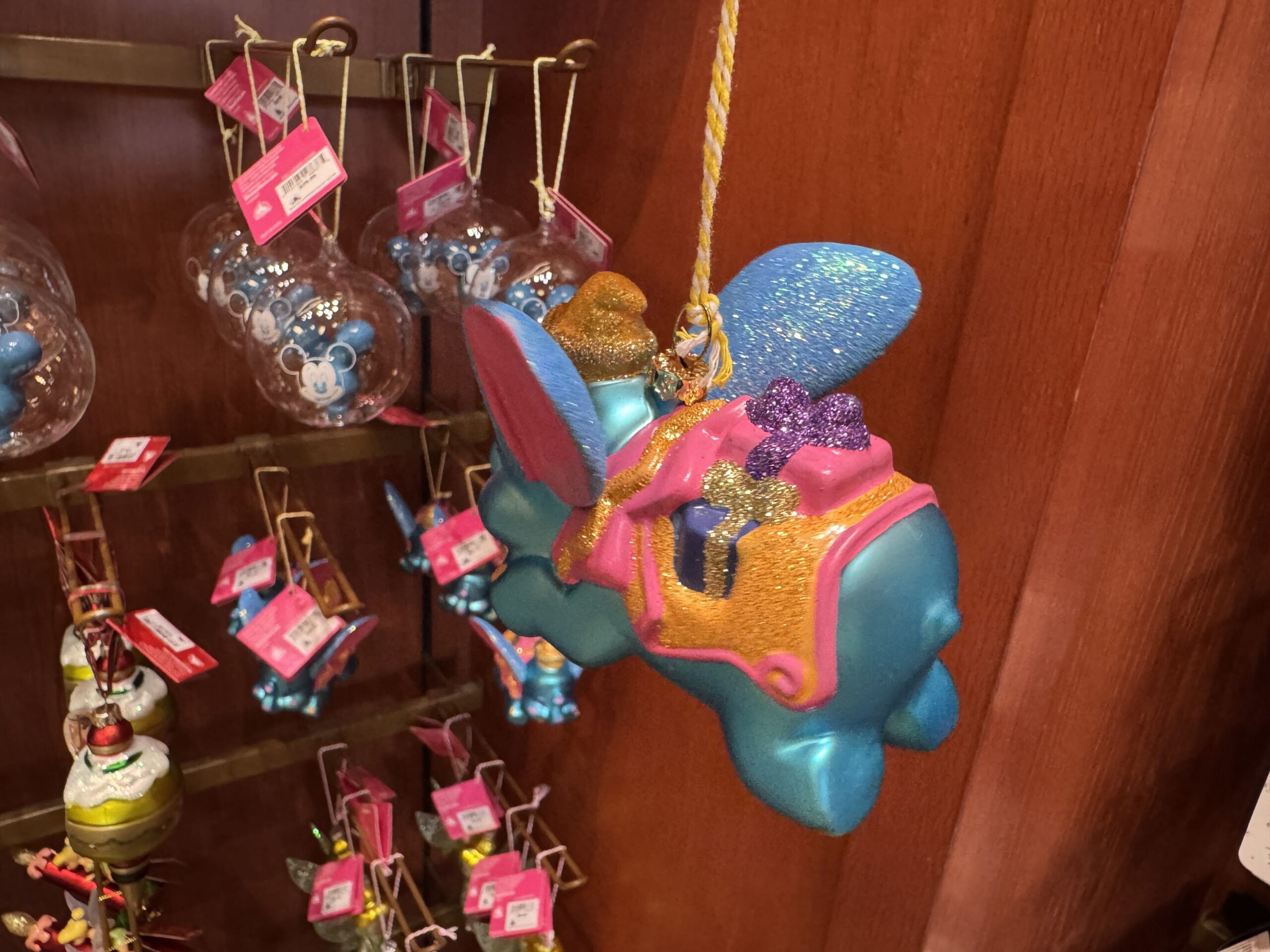 Dumbo flies in bright, cheery colors of blue, orange, and pink. His ears, hat, and saddle holding presents sparkle with glitter. He is crafted of porcelain and painted with rosy cheeks in a watercolor style. 