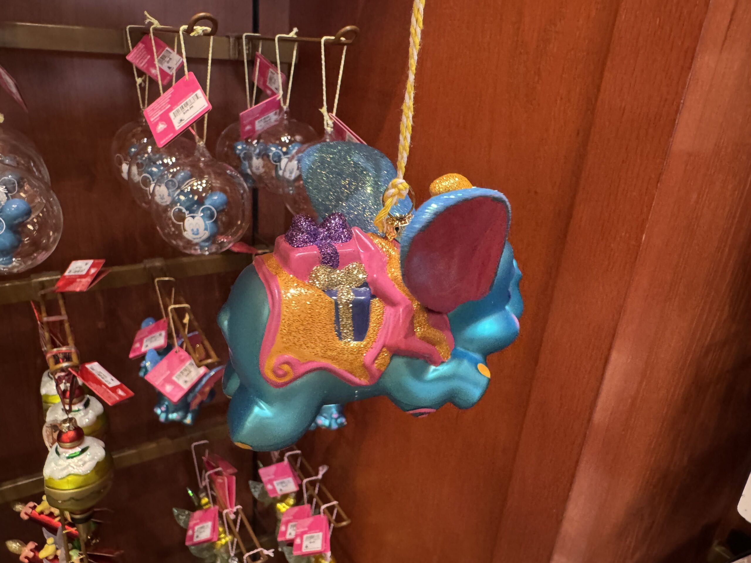 Dumbo flies in bright, cheery colors of blue, orange, and pink. His ears, hat, and saddle holding presents sparkle with glitter. He is crafted of porcelain and painted with rosy cheeks in a watercolor style. 