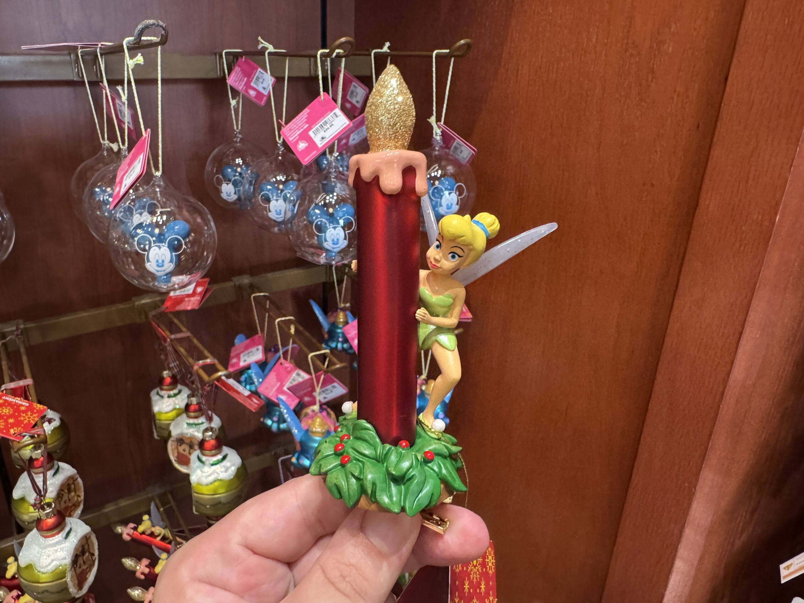 Here we find Tinker Bell peeking out from behind a red candle with a glittery flame, wearing a sassy smile. Her wings are frosted with sparkles of glitter pixie dust. Both stand atop a tuft of holly and red berries. 