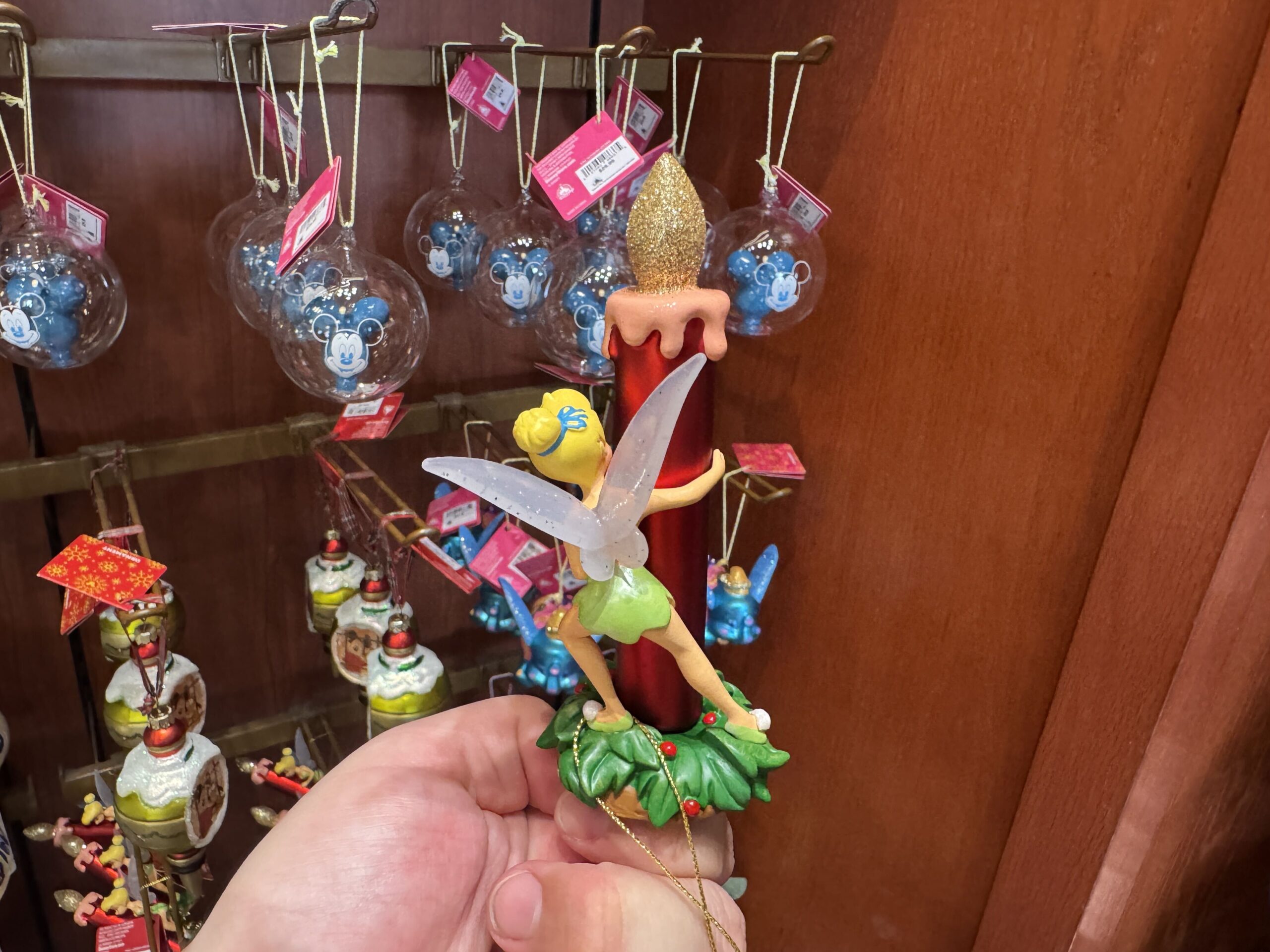 Here we find Tinker Bell peeking out from behind a red candle with a glittery flame, wearing a sassy smile. Her wings are frosted with sparkles of glitter pixie dust. Both stand atop a tuft of holly and red berries. 