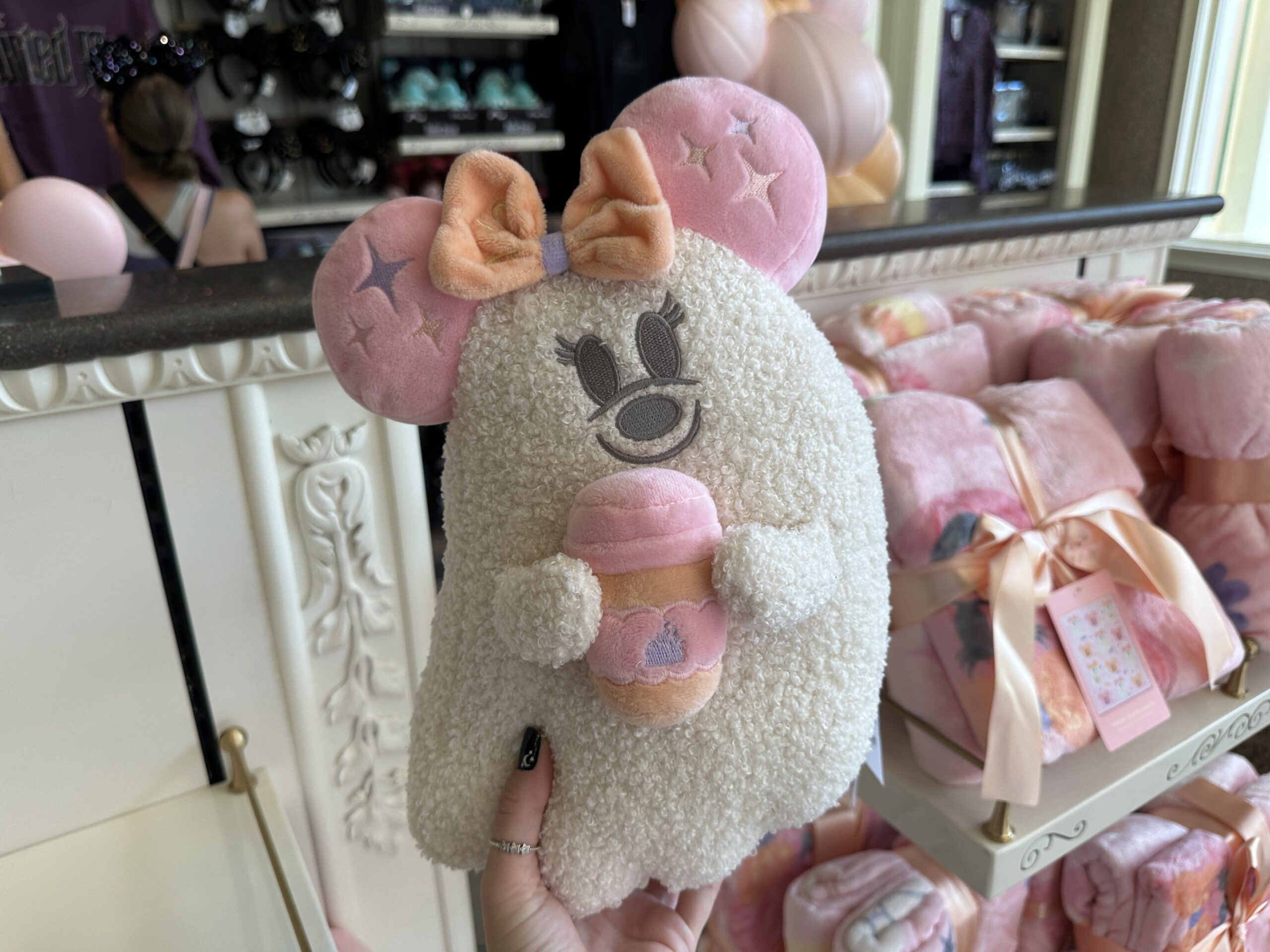 Also in the collection is a Minnie ghost-shaped pillow. It's made out of sherpa. She carries a coffee cup in her hands as she wears a beige and purple bow. Her ears are pink.