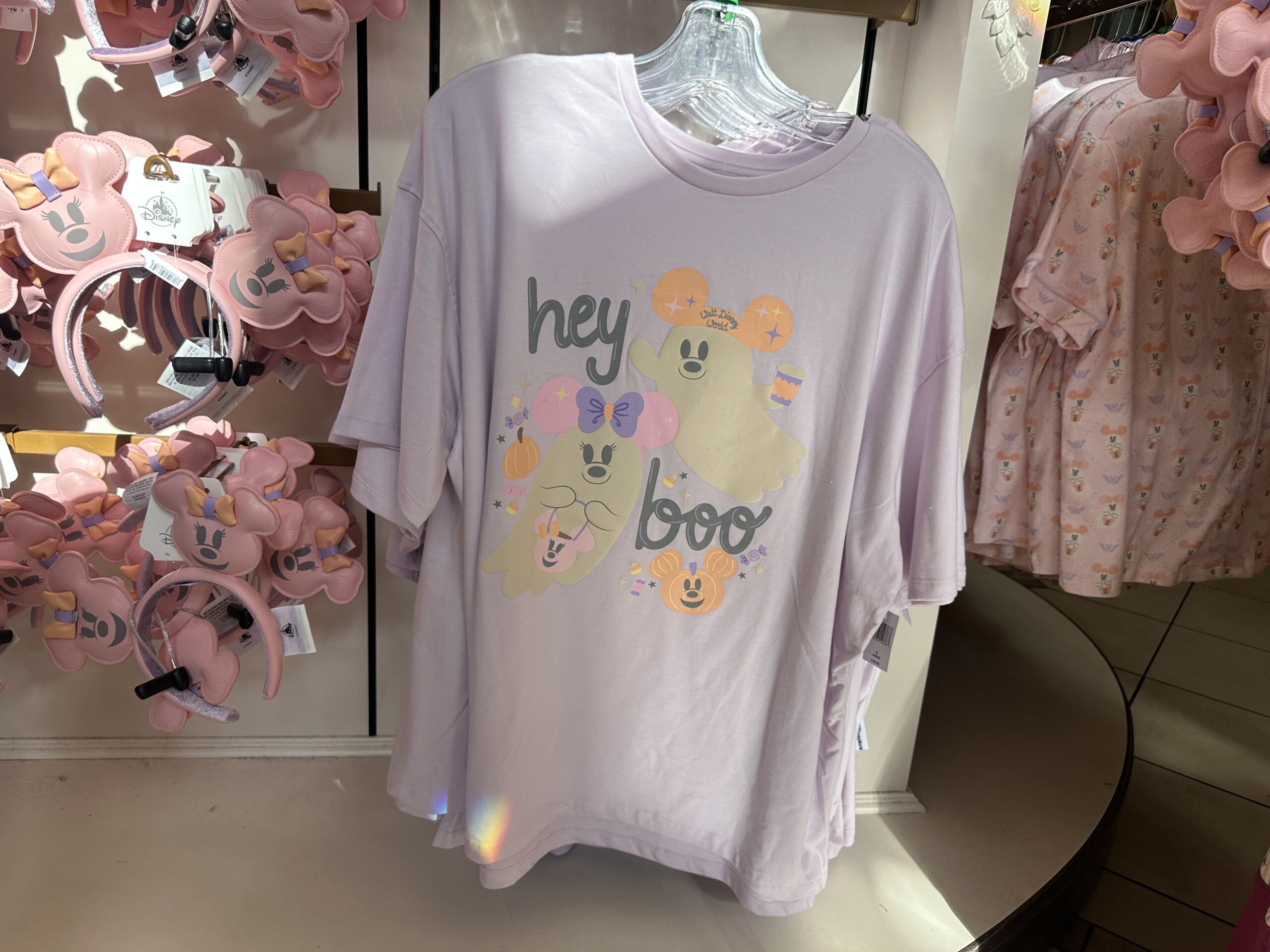 The purple T-shirt has Mickey and Minnie ghosts on the front. Mickey holds a coffee cup, and Minnie holds a candy bucket. More jack-o'-lanterns and candy surround them. "hey boo" is in a light gray script.