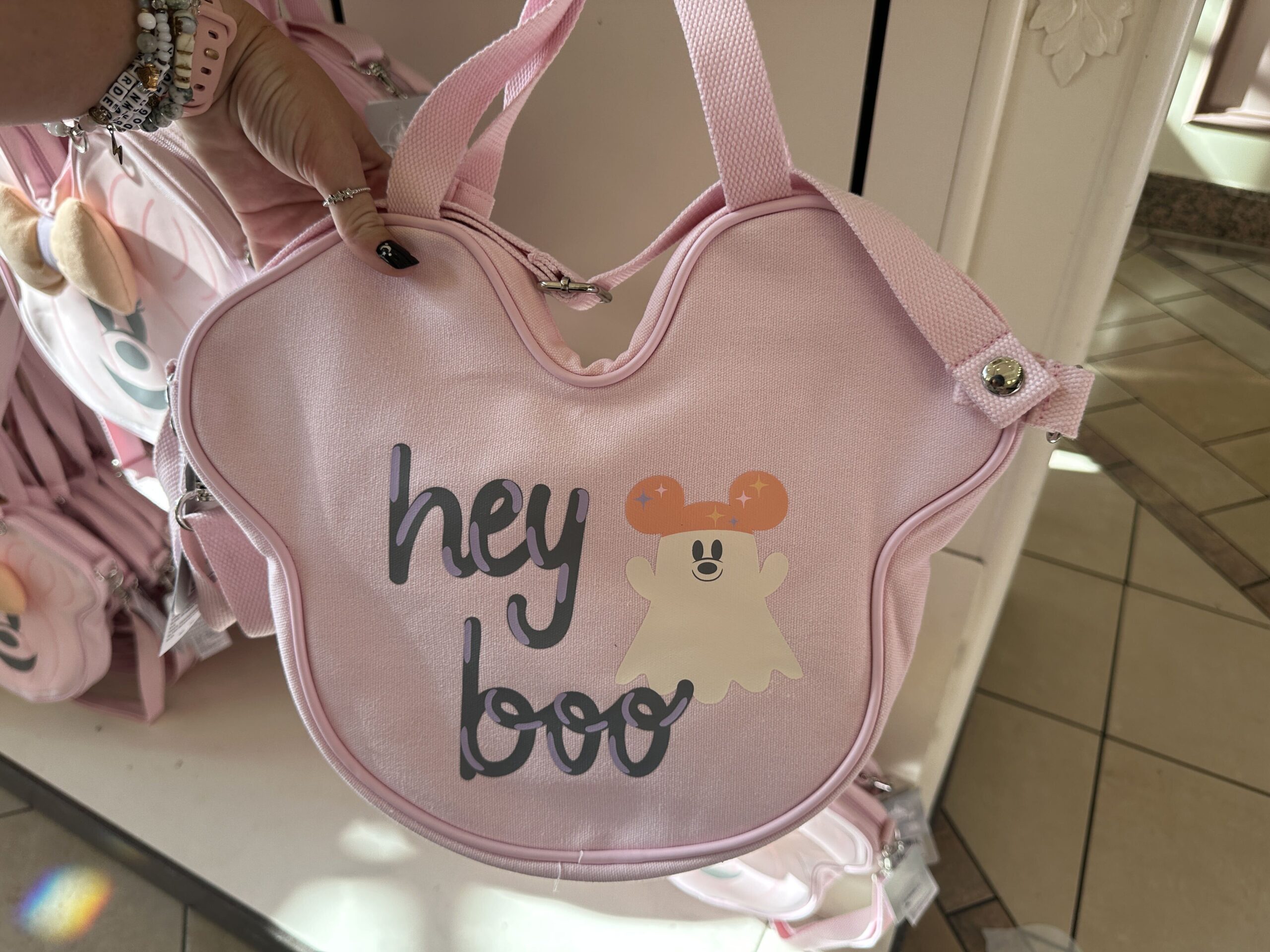 A pastel pink face tote is part of the collection. Shaped like a jack-o'-lantern, Minnie has a three-dimensional orange and purple bow. The back say "hey boo" and has a Mickey ghost. It has a shoulder strap, as well as a secondary strap that can be removed.