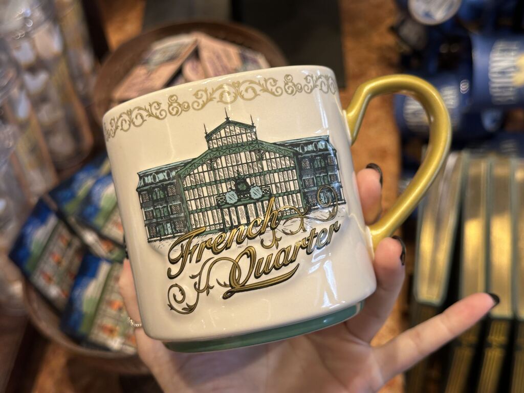 On the other side, the exterior of the main entrance is shown in green, black, and blue. "French Quarter" is seen right below it in black and gold cursive writing. The outline of the bottom of the mug is green.