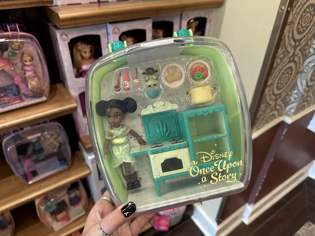 Meanwhile, Tiana's case features a miniature doll wearing a green patterned dress, stove, kitchen utensils, fake food, and two shelves to store the small items. The set also features a small doll wearing a green dress and a bow.